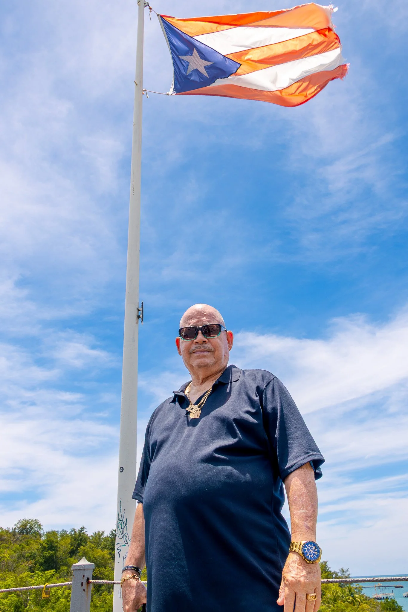 A man wearing sunglasses, a gold watch, and jewelry stands outdoors under a blue sky with wispy clouds. Behind him is a flagpole with a worn semi-tattered Puerto Rican flag.