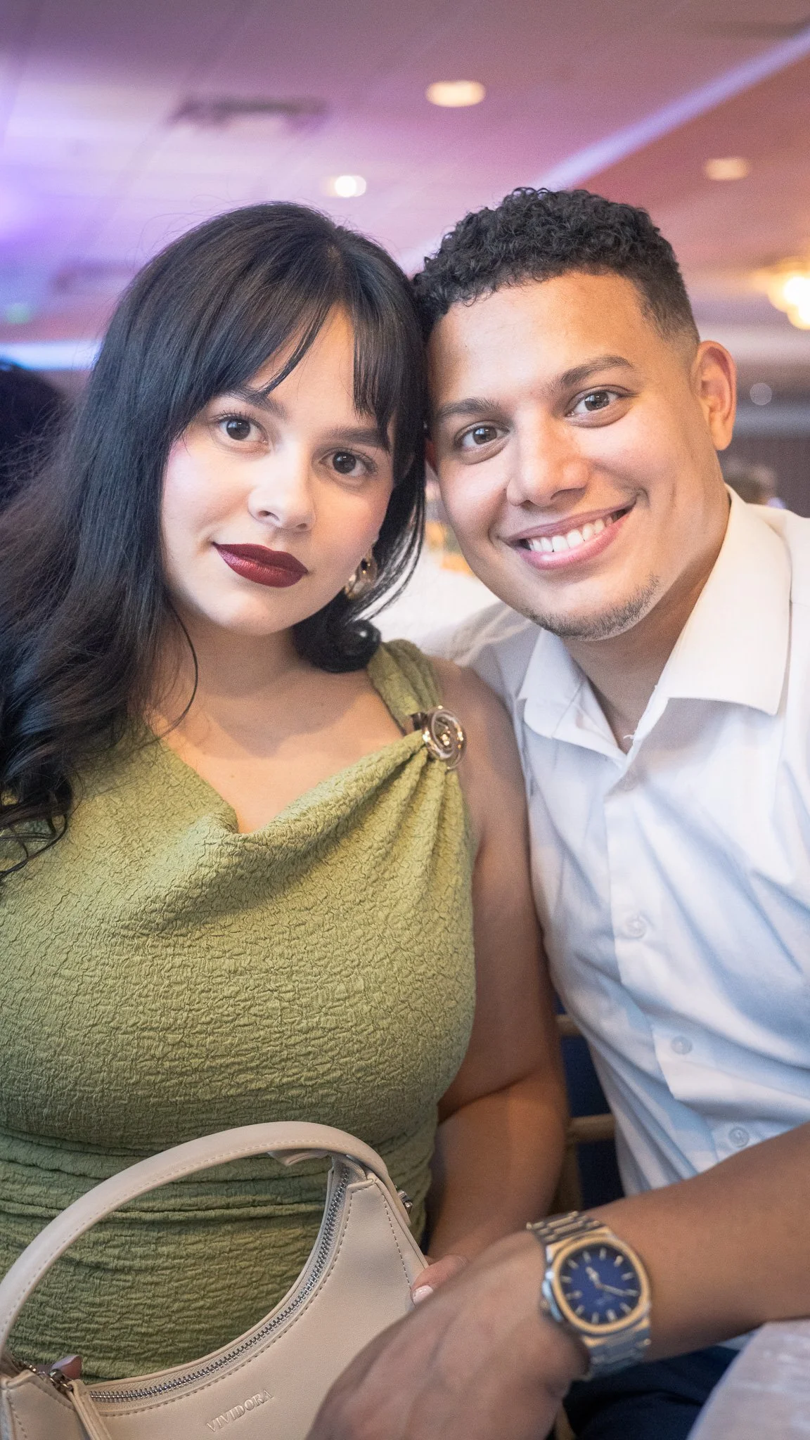 A smiling woman with dark hair and red lipstick sitting next to a smiling man with short curly hair in a white shirt. They are in a dimly lit room with purple and warm lighting, possibly at an event or restaurant.
