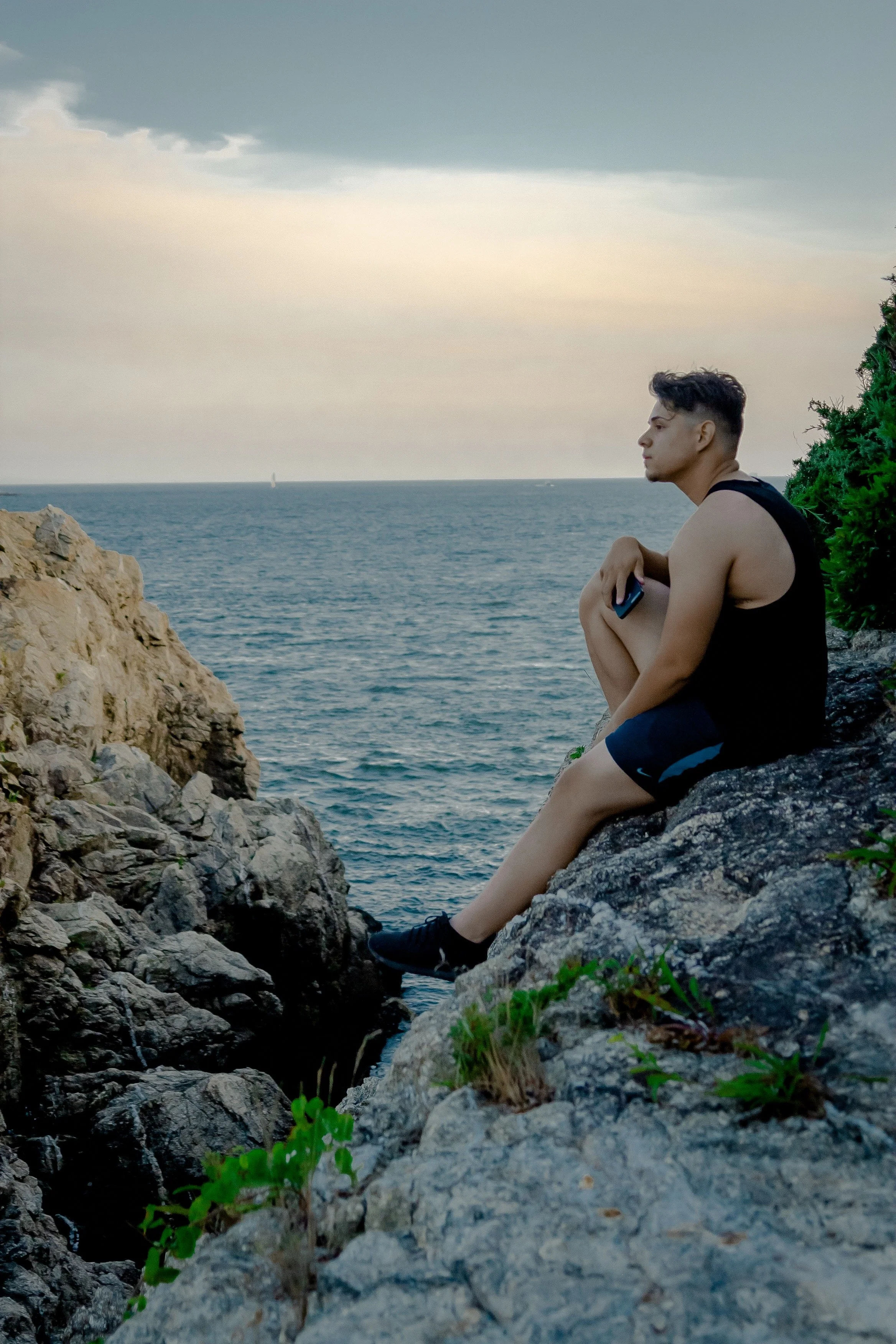A young man in a black tank top and shorts sits on rocks by the ocean, holding a phone, with a cloudy sky overhead.
