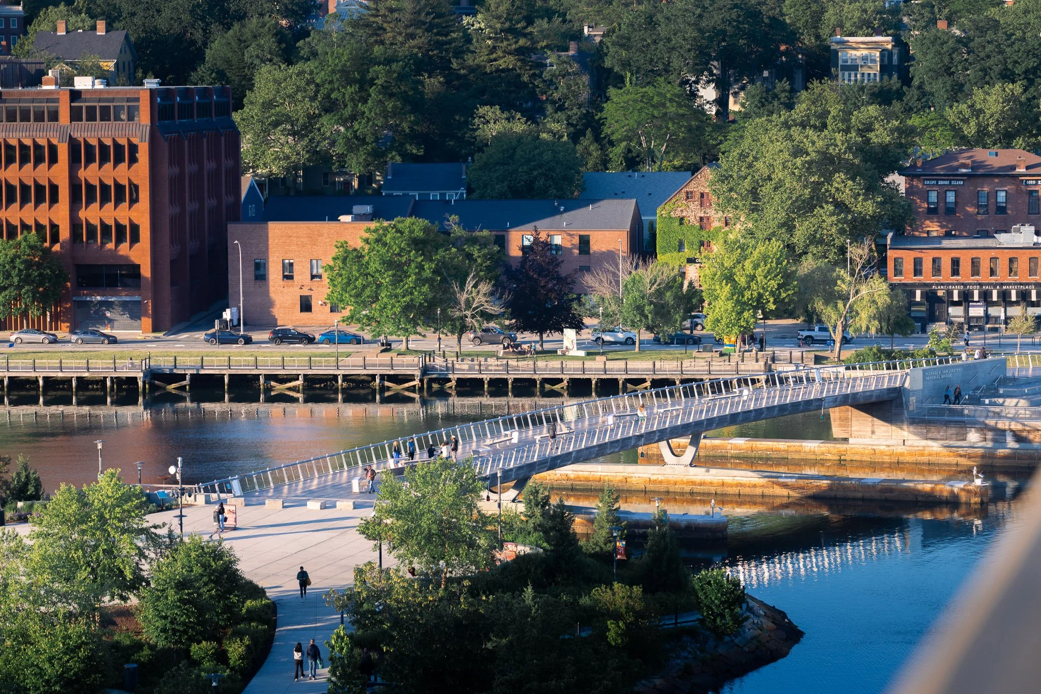 An aerial view of a cityscape featuring various buildings, green trees, a river, and a pedestrian bridge with people walking on it.