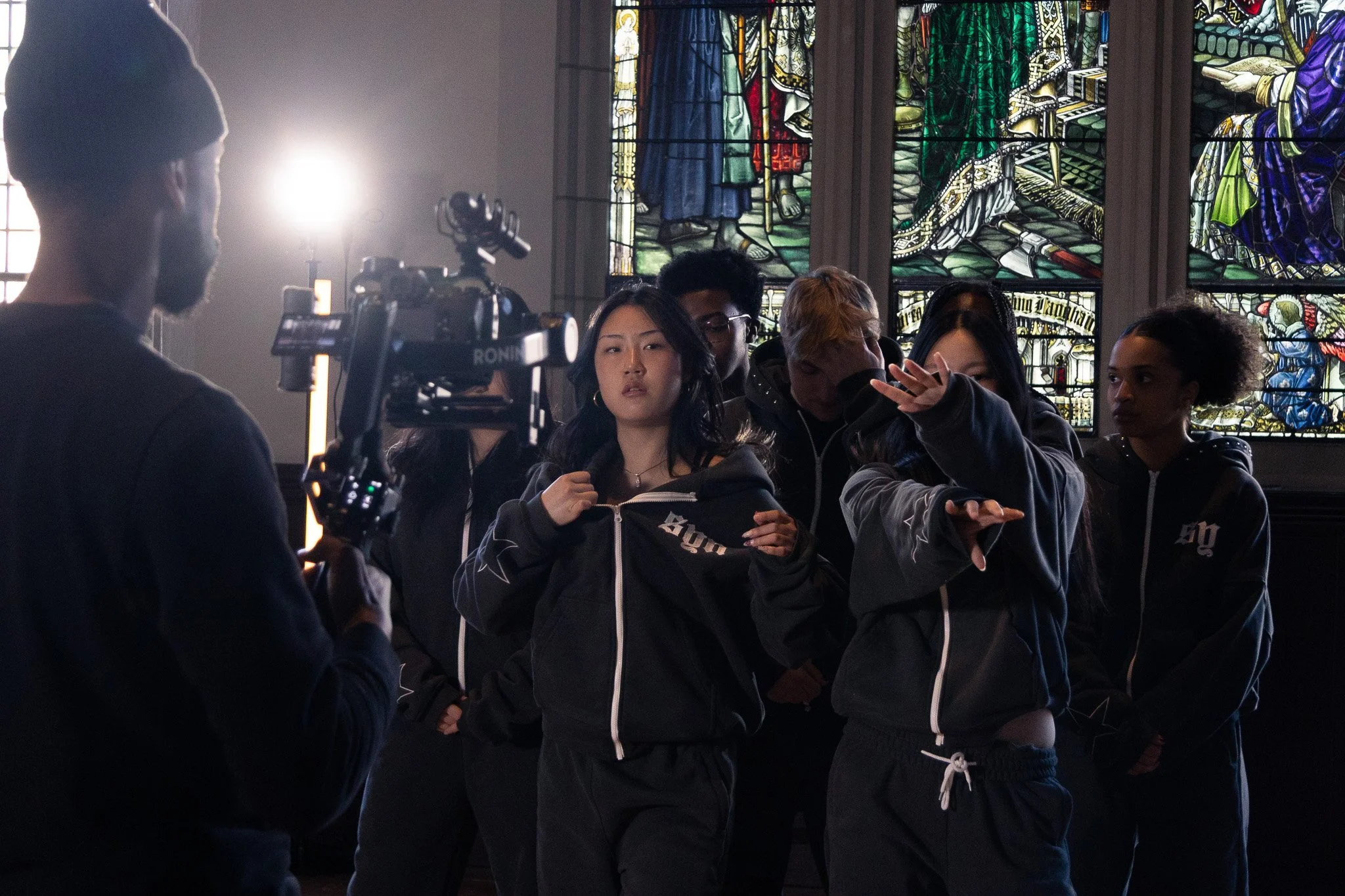 A group of young people standing inside a church with stained glass windows in the background. They are dressed in dark clothing, some with hoodies, and appear to be listening or waiting, with some looking towards a person on the left who is holding 