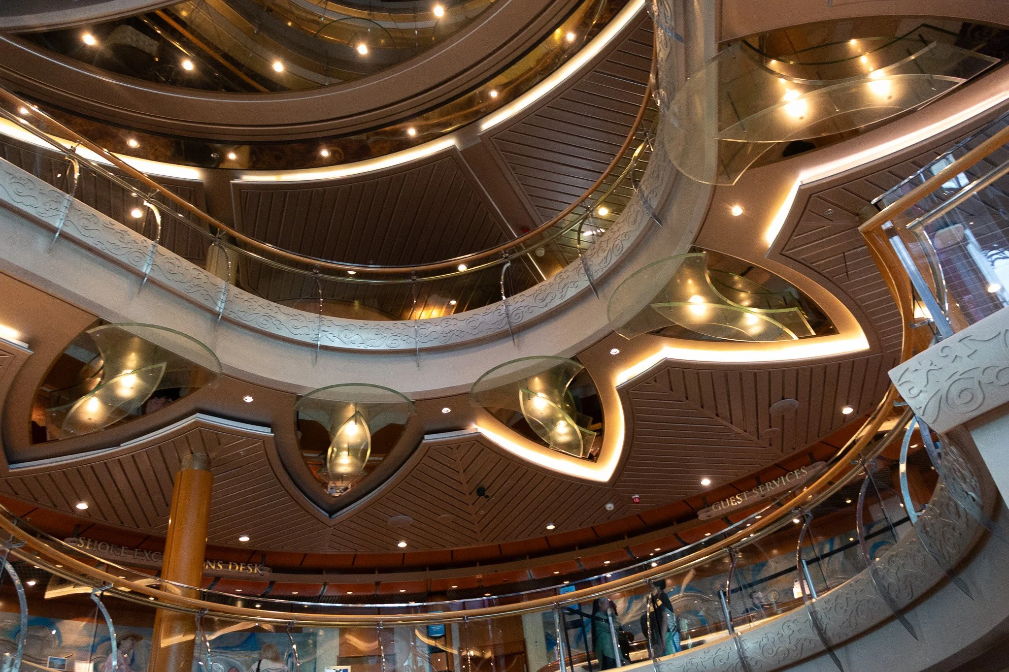 Multiple floors inside a modern building with curved, metallic and glass railings, warm lighting, and a decorative ceiling design.