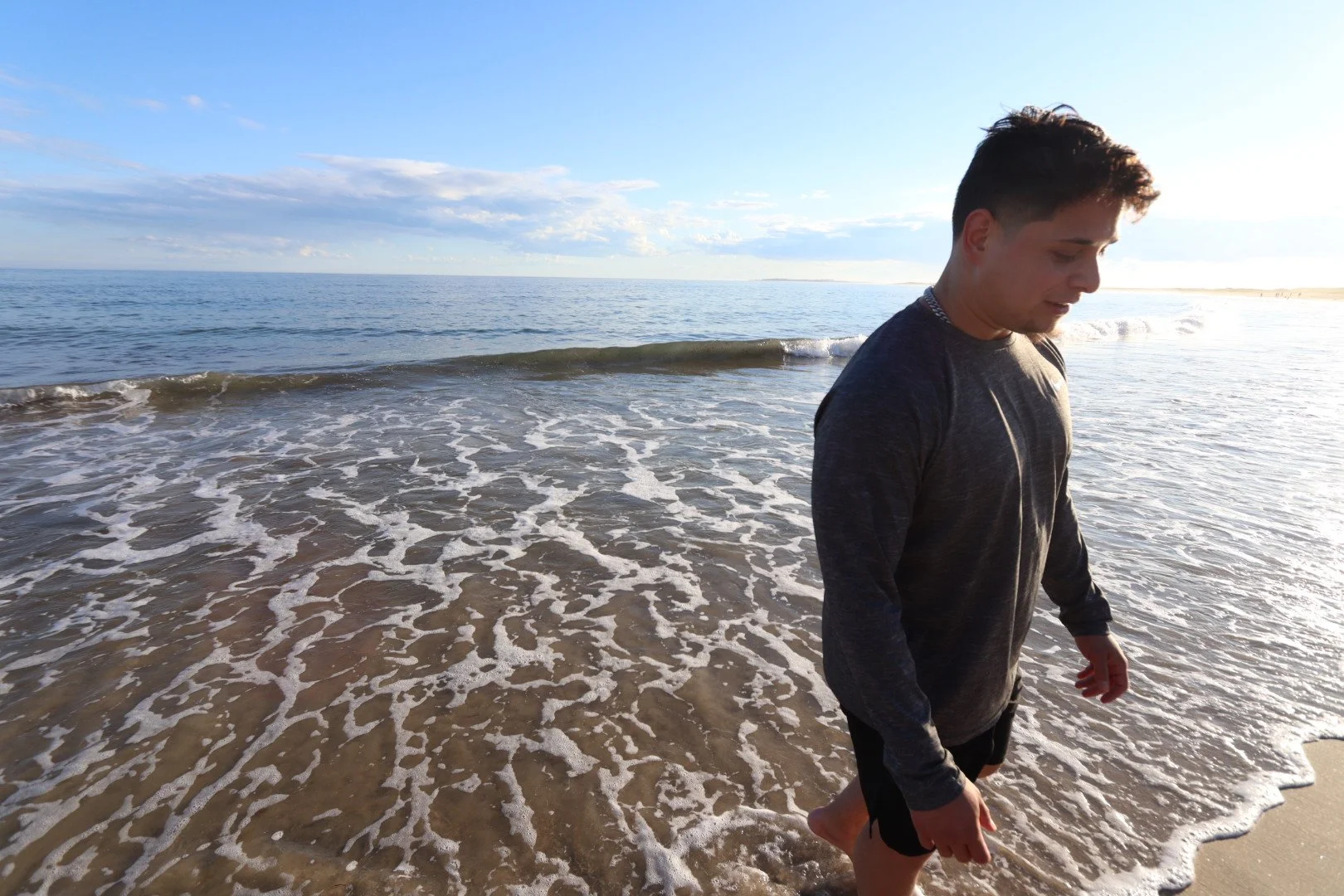 A young man with short dark hair walks along the shoreline with gentle waves and sunlight on the water, wearing a long-sleeve dark shirt and shorts.