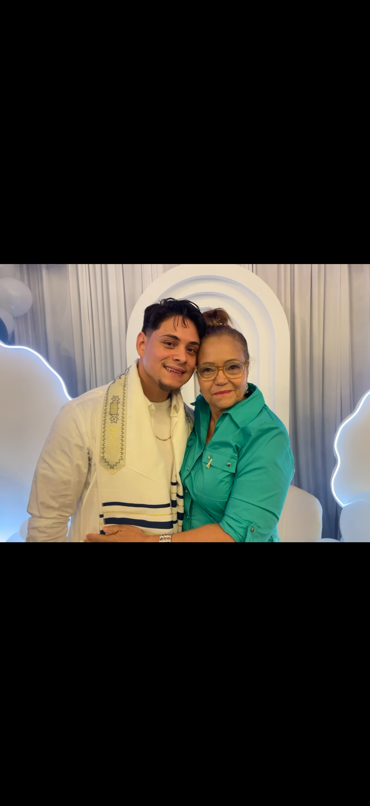 A young man and an elderly woman with glasses, smiling and hugging, in front of a decorative backdrop.
