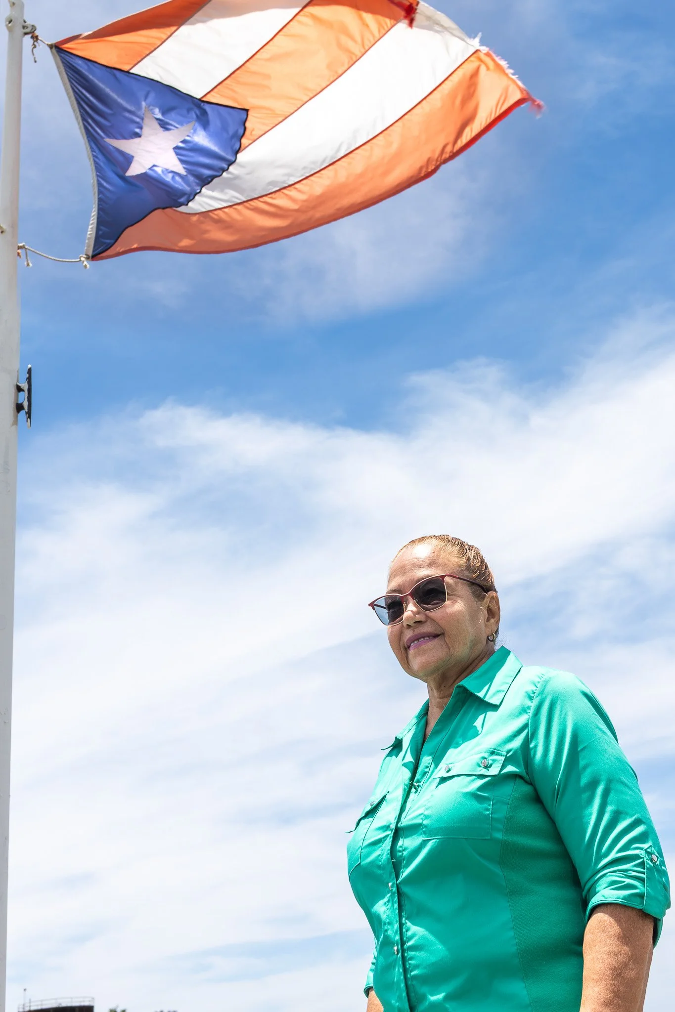 A woman in sunglasses and a teal shirt walking outdoors on a sunny day with a partly cloudy sky. An American flag is flying in the background.