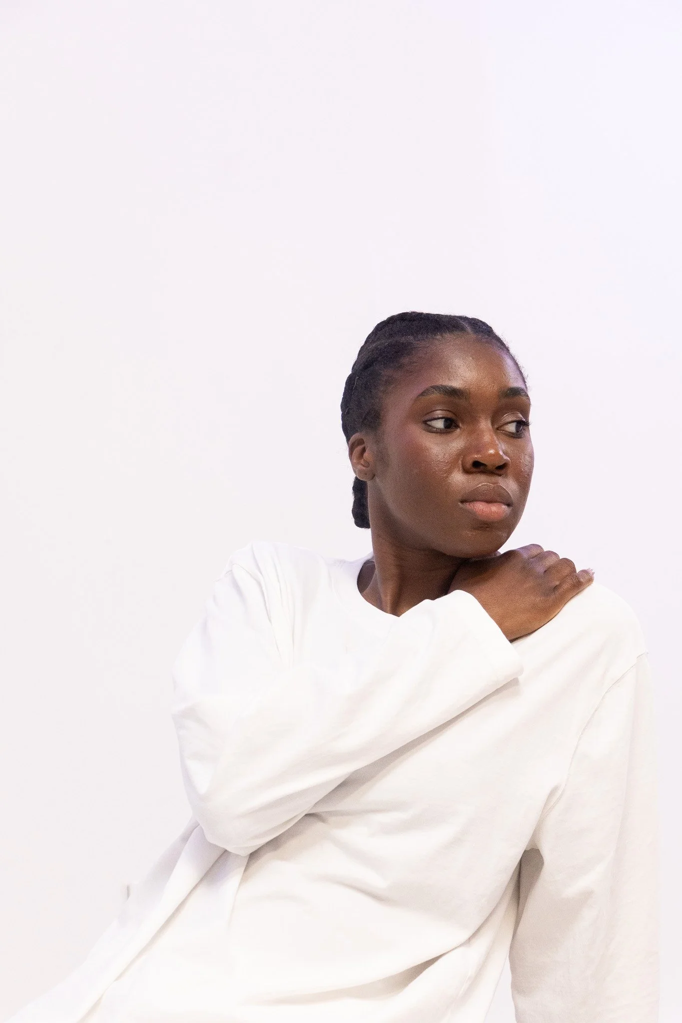 A woman with braided hair and dark skin wearing a white long-sleeve top, sitting against a plain white background, resting her chin on her crossed arms.