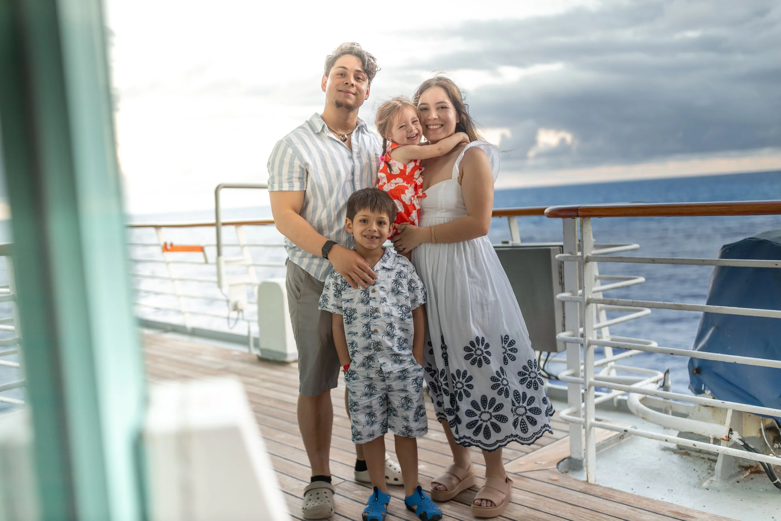 A family of four, including a woman, a man, and two children, standing on a boat deck near the ocean, smiling and hugging each other with a cloudy sky in the background.