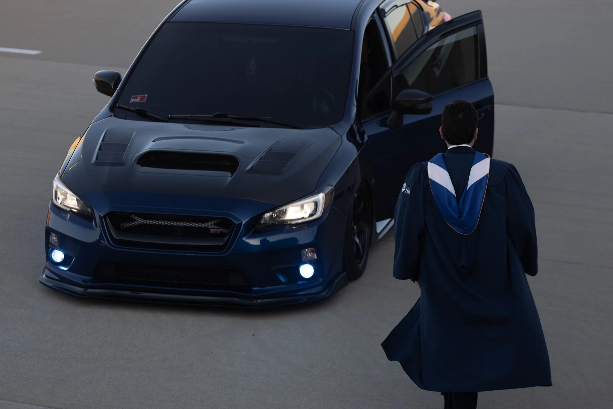 A person in graduation attire standing next to a dark blue car in a parking lot.