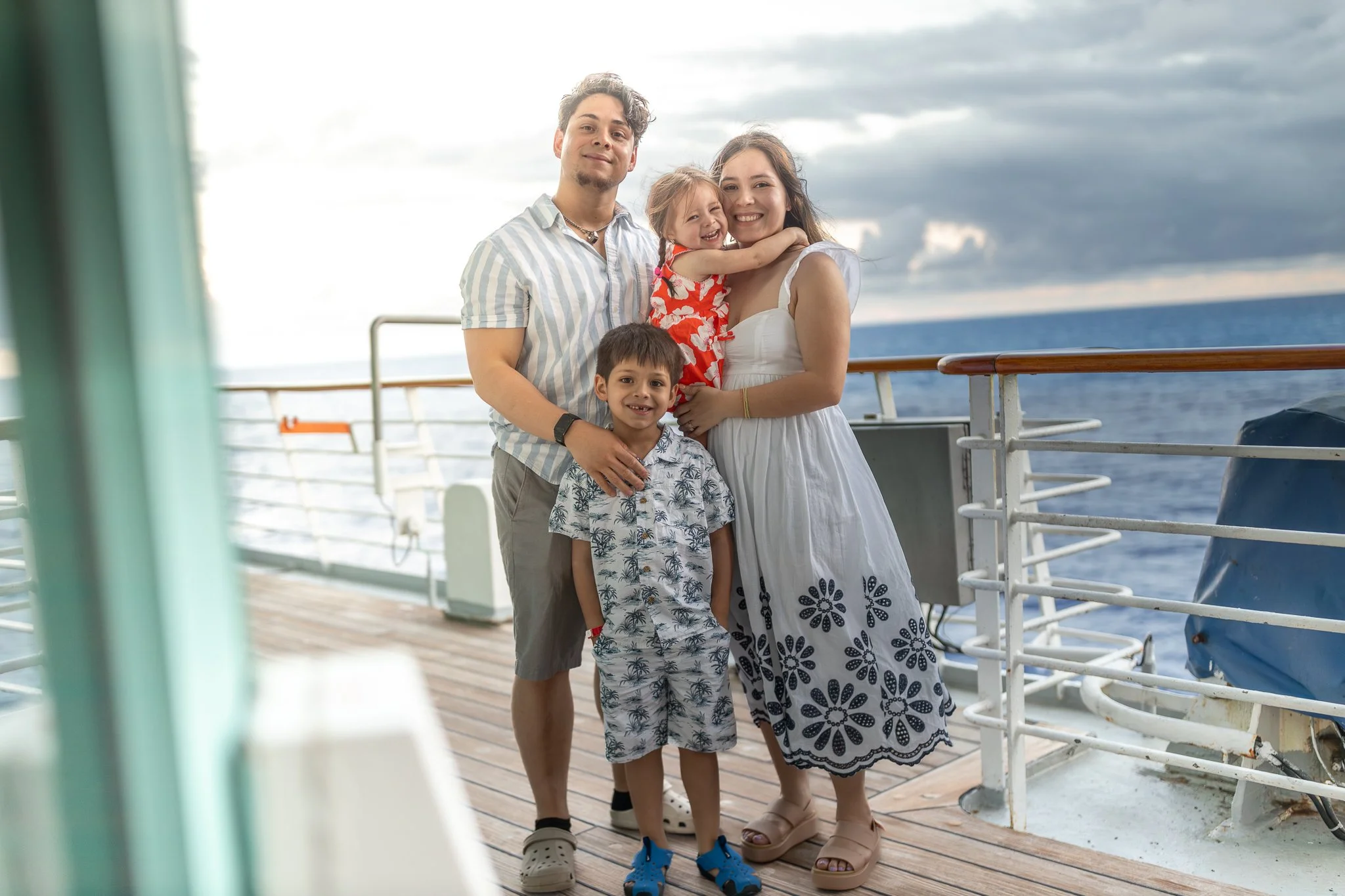 Family of four on a ship's deck with the ocean and cloudy sky behind them. The mother and two children are smiling, the father stands beside them. The children are a girl with a red floral dress and a boy with a tropical print shirt. The family appears to be enjoying their time at sea.
