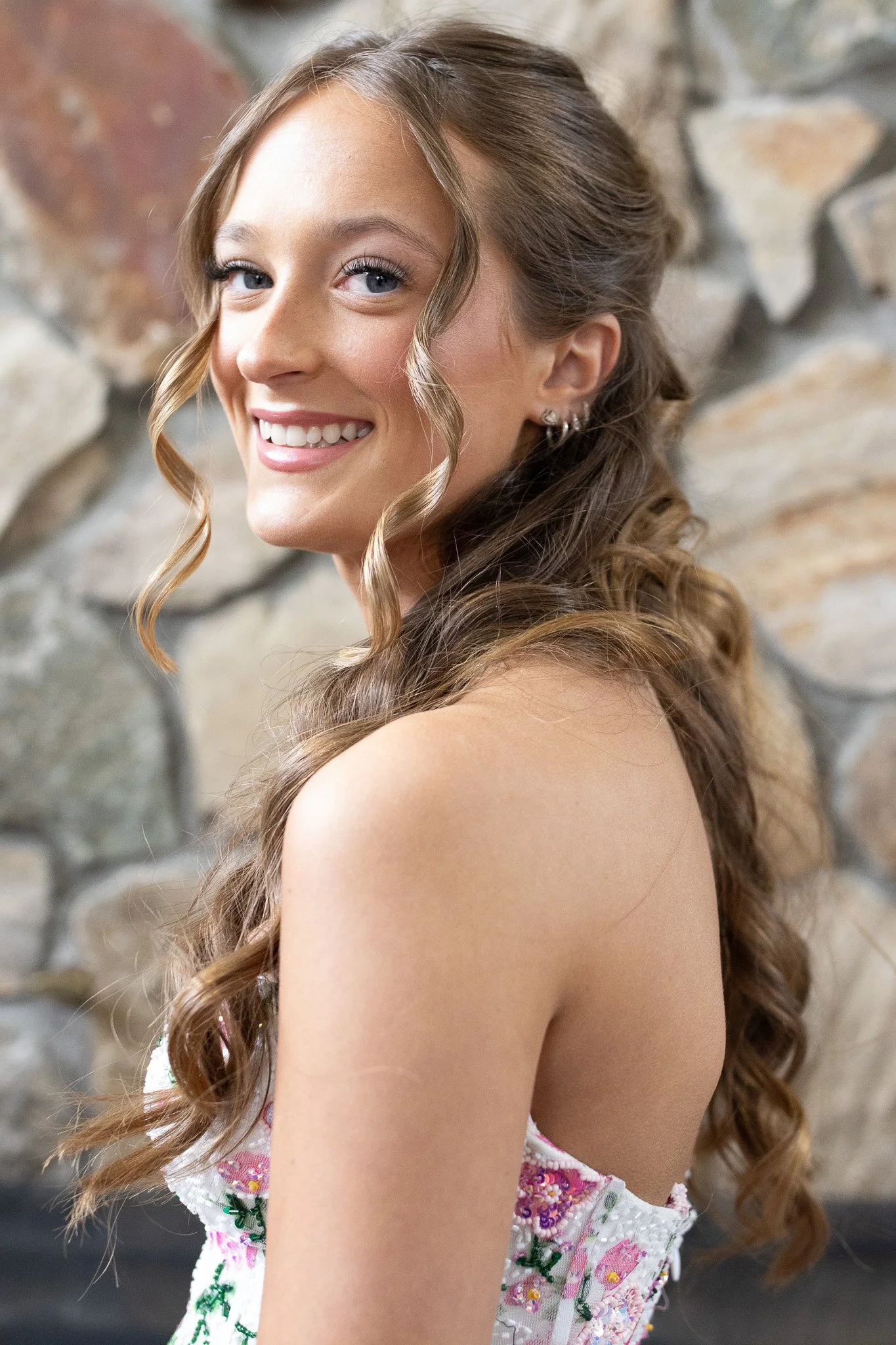 Young girl with long curly brown hair styled in loose curls, smiling, wearing a sleeveless floral dress with pink, green, and white colors, a stone wall in the background.