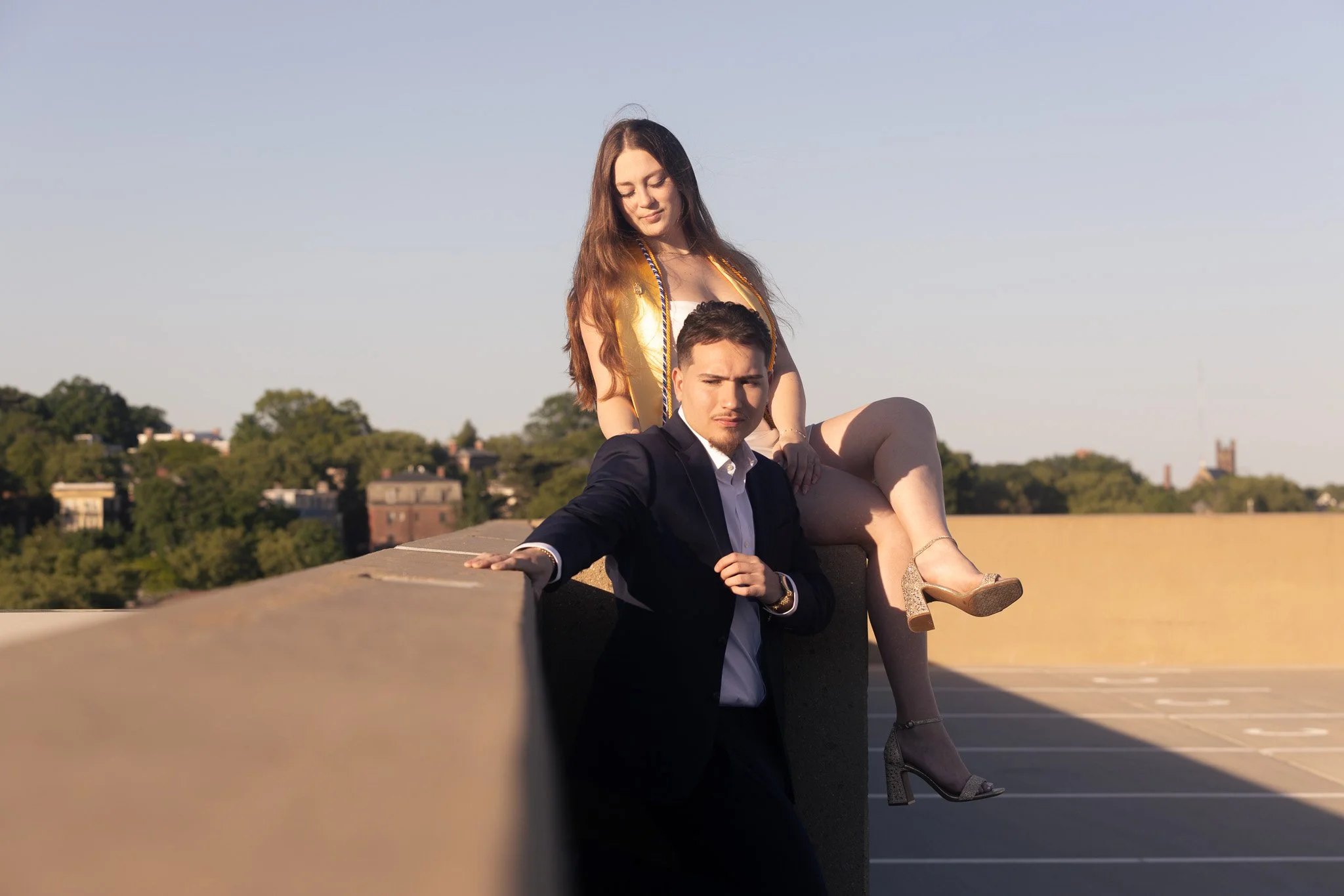 A young woman with long brown hair, wearing a white dress and gold high heels, sitting on a ledge with her eyes closed, atop a parking garage. A young man in a suit with a white shirt and black jacket, sitting nearby with one hand on the ledge, looki