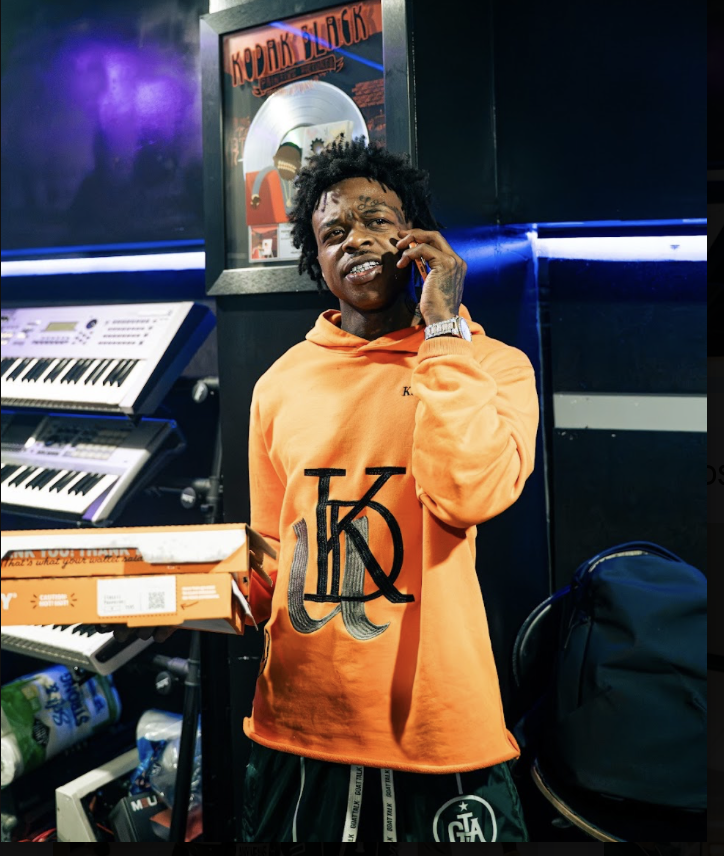 A young man with curly hair and tattoos, wearing an orange hoodie, is holding a phone to his ear and a pizza box in a music studio or entertainment space. There are keyboard instruments and a framed platinum record on the wall behind him.