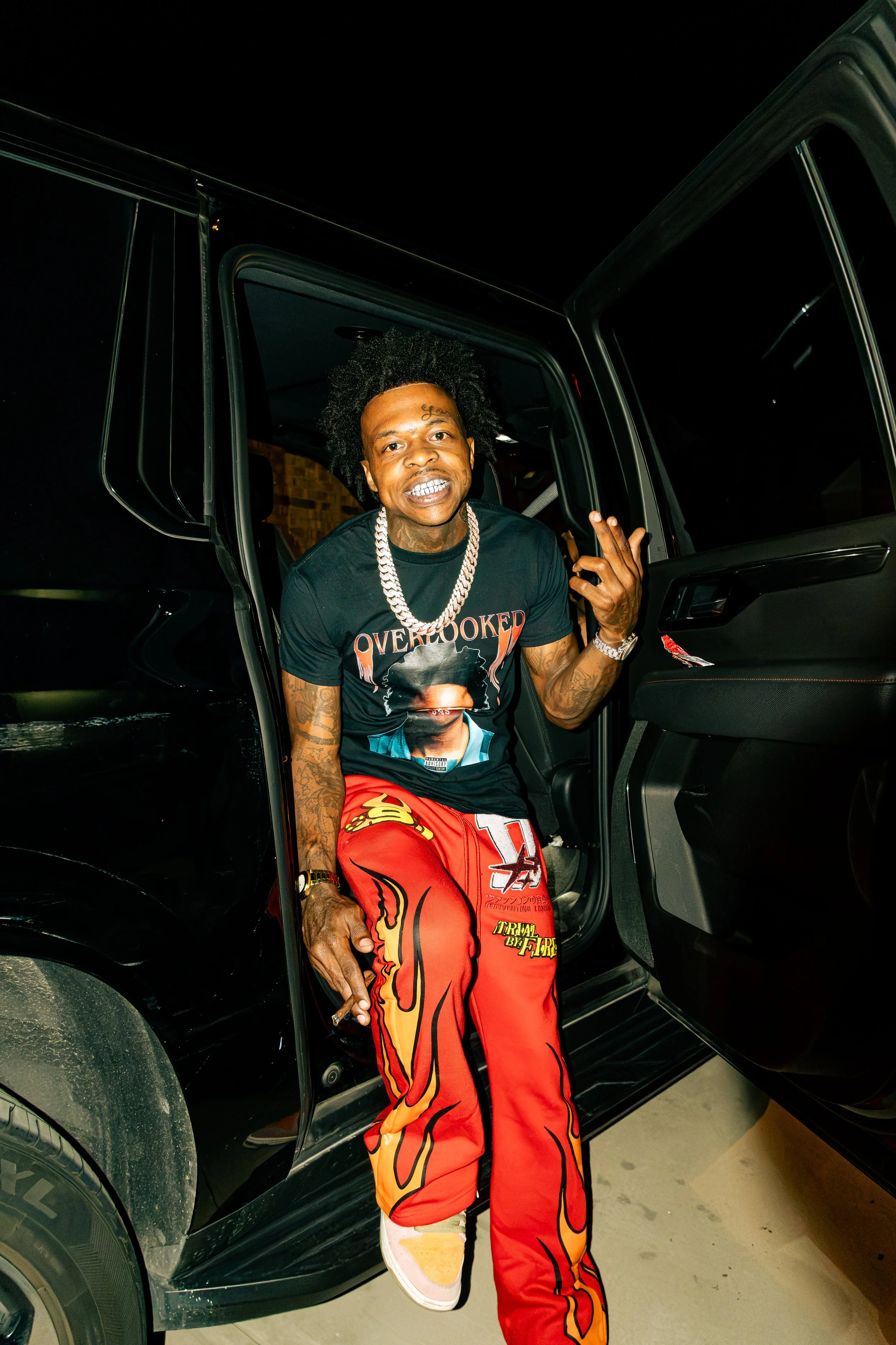 A man with curly hair and tattoos on his arms sitting in a black car at night, wearing a black T-shirt, red and orange flame-patterned pants, a large chain, and holding a cigarette.