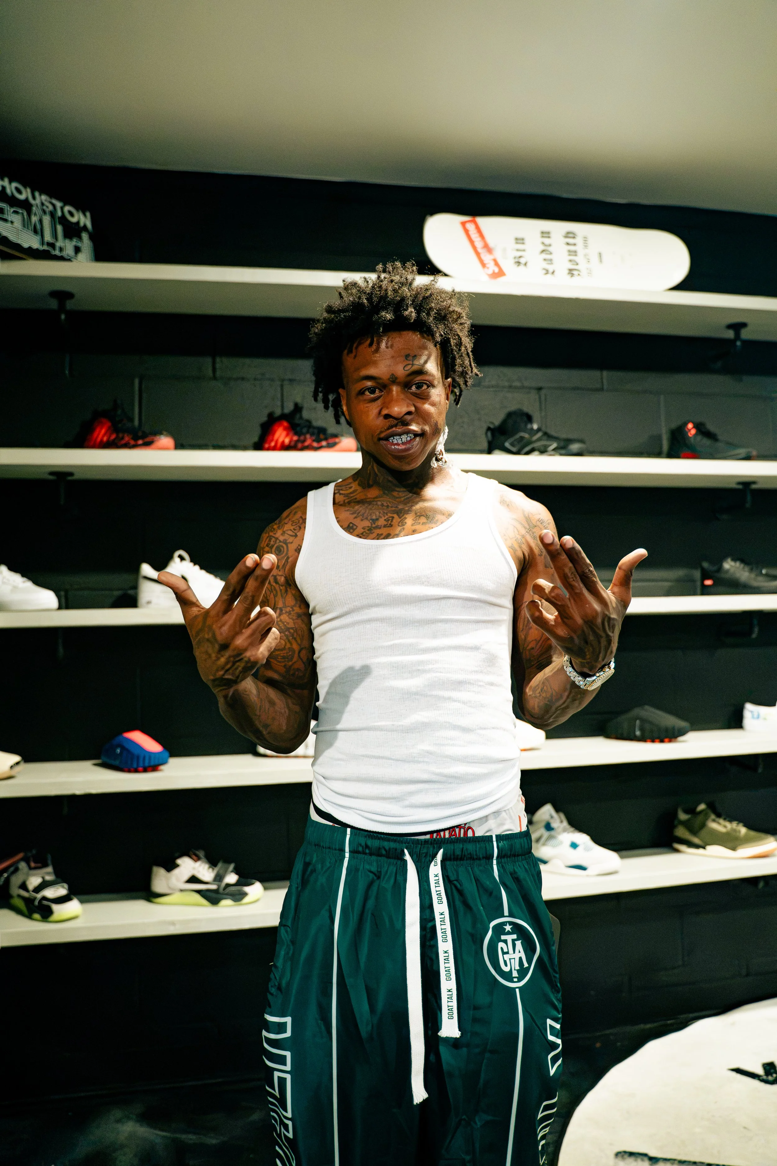 A young man with tattoos on his arms and chest, wearing a white tank top and green shorts with white drawstrings, standing in a sneaker store with shelves of sneakers in the background and a skateboard on top of the shelf.