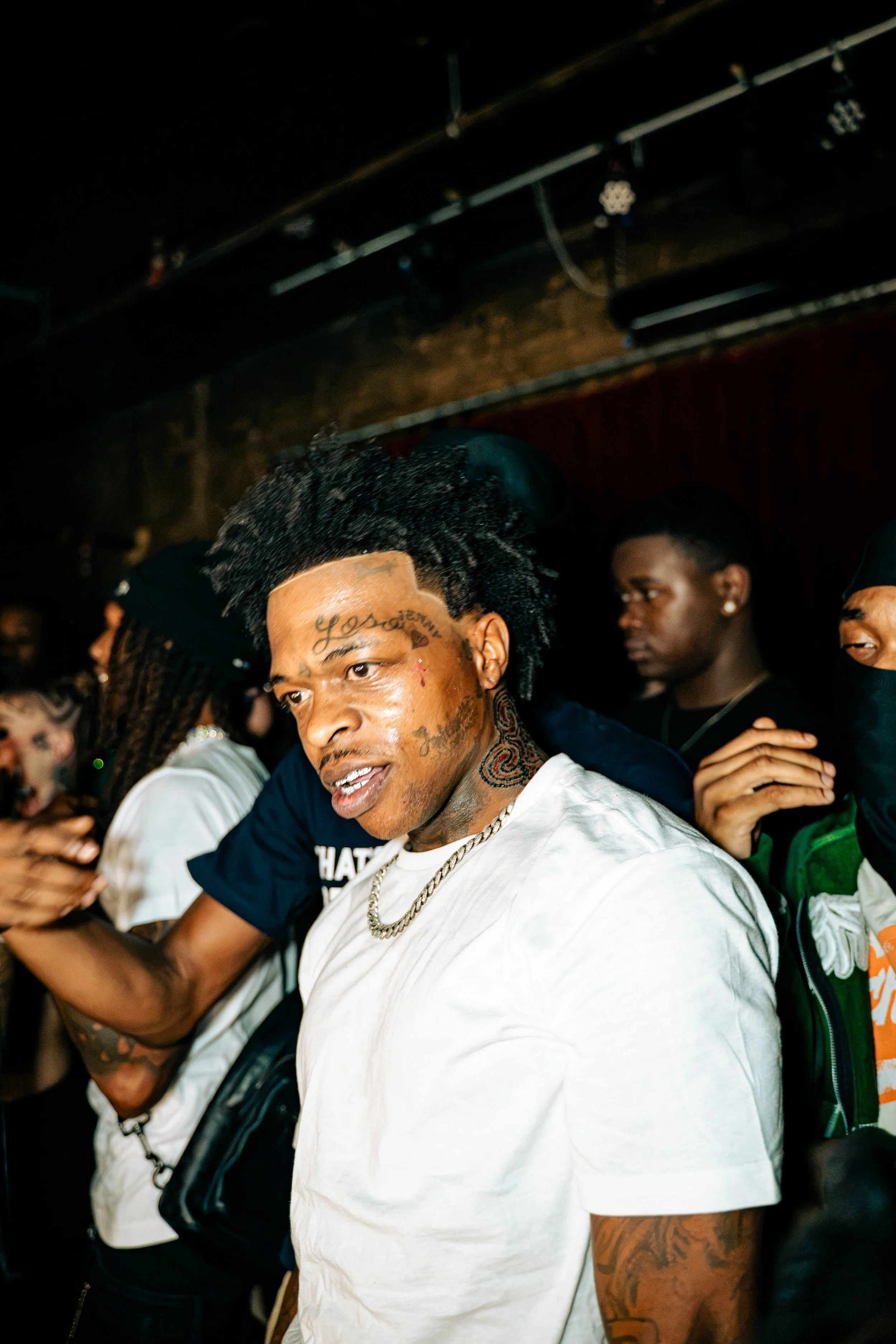 A group of people at a party or nightclub, with focus on a man with tattoos, dreadlocks, and earrings, wearing a white T-shirt and a silver chain, surrounded by others
