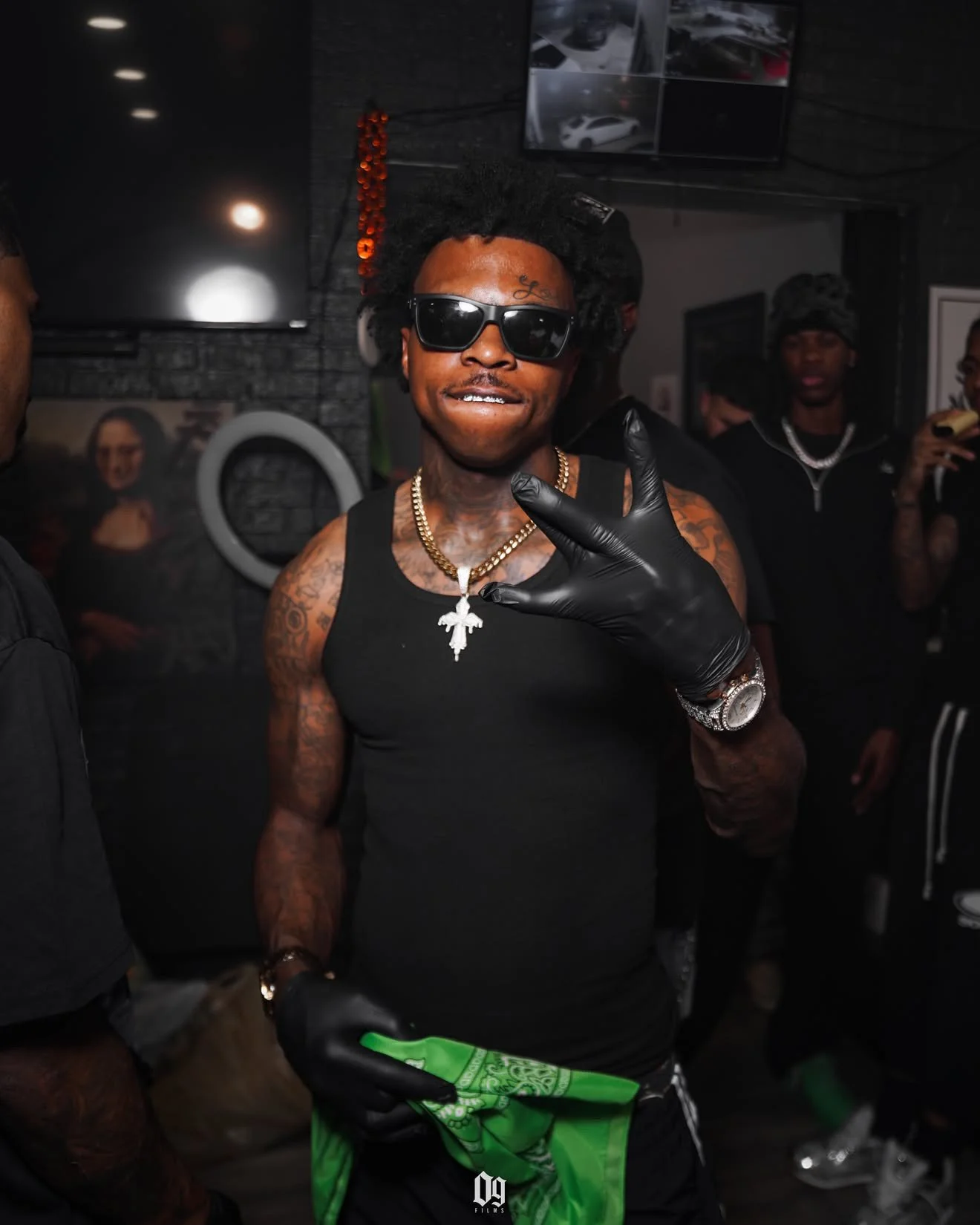 A man with dark sunglasses, gold chains, and tattoos making a hand gesture, wearing a black sleeveless shirt, black gloves, and holding green money, in a dimly lit indoor setting with other people in the background.