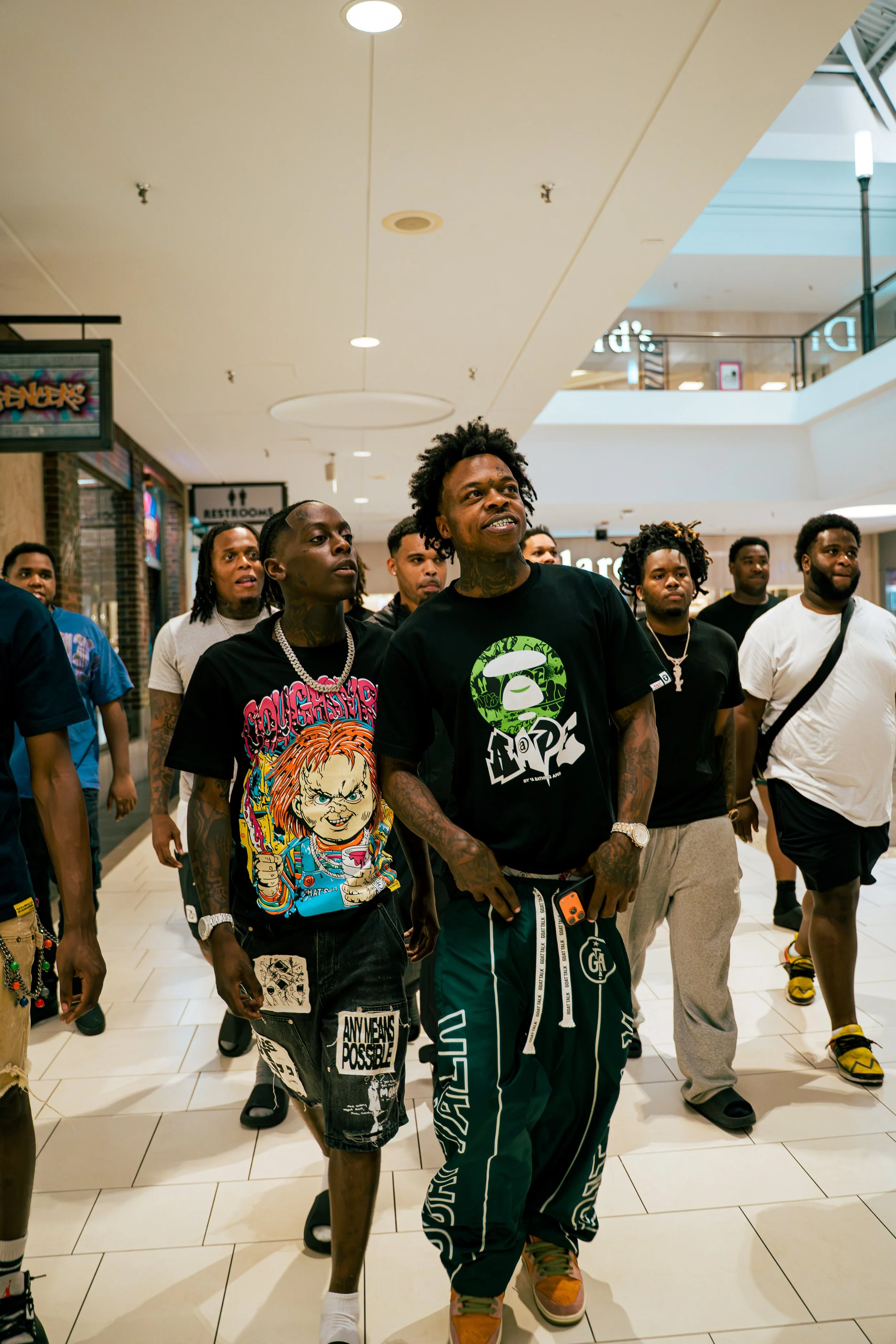 A group of young men walking in a shopping mall, wearing casual streetwear and jewelry, with some tattoos visible on their arms.