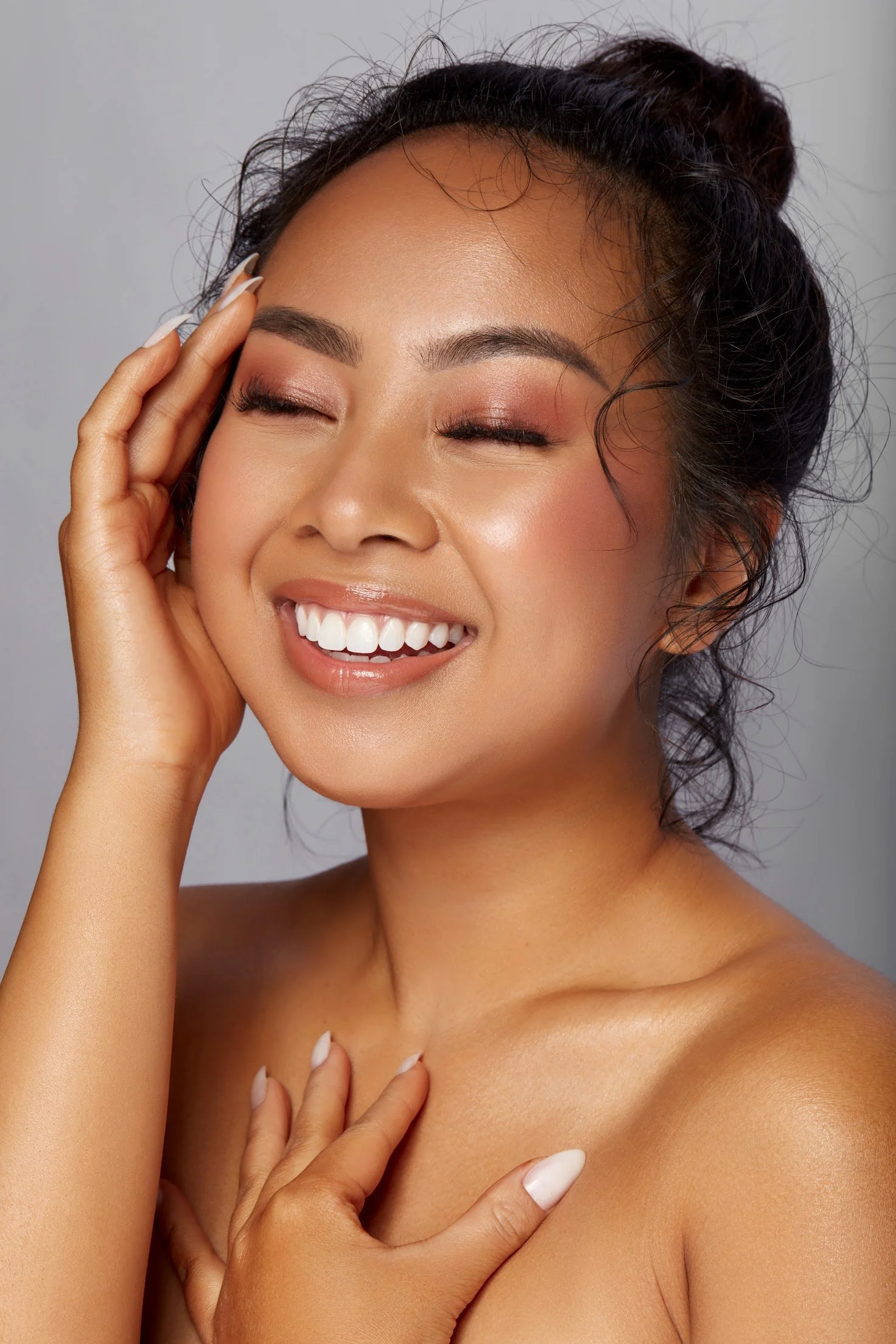 Close-up of a smiling woman with closed eyes, touching her face and chest, with natural makeup and wavy hair.