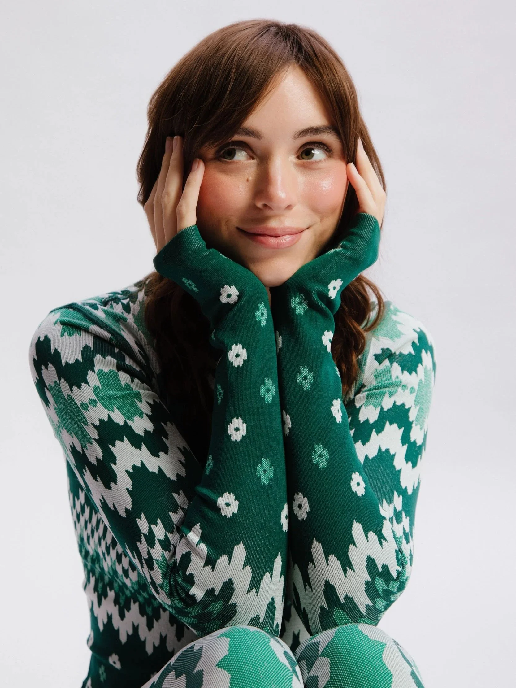A young woman with brown hair styled in loose waves, touching her face with both hands, wearing a green sweater with white and dark blue patterns, and patterned pants with green, white, and turquoise colors, sitting against a plain white background.