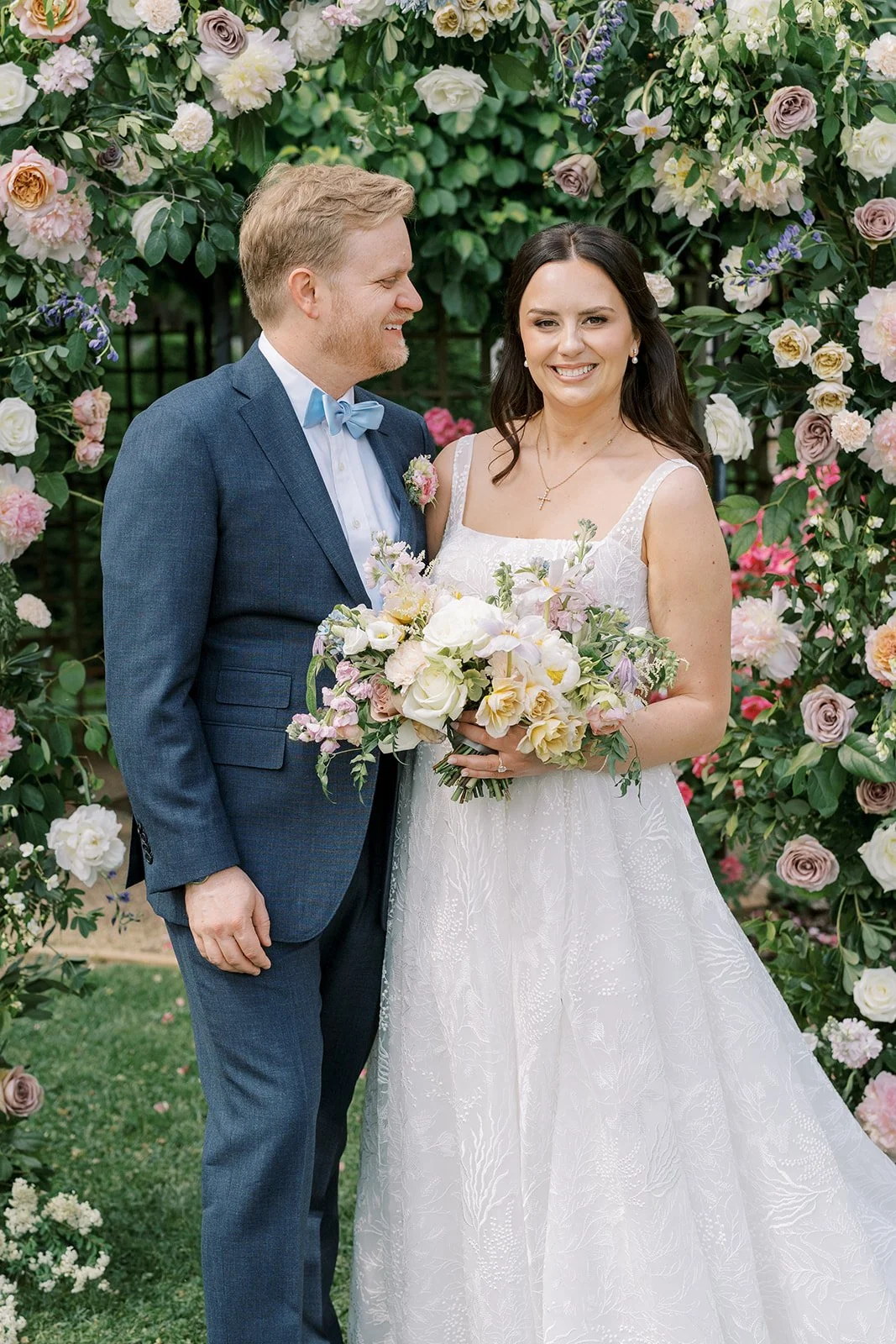 A bride and groom pose together in a garden with a floral arch, smiling. The bride wears a white wedding dress and holds a bouquet of flowers, while the groom wears a blue suit with a light blue bow tie.