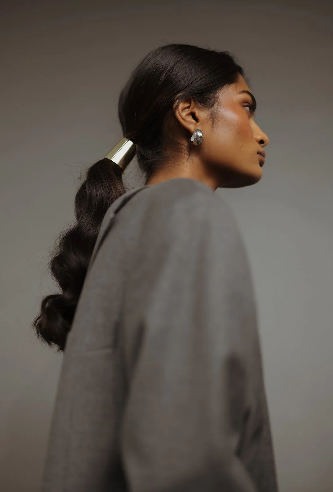 Side profile of a woman with long, dark wavy hair tied in a ponytail with a metallic hair accessory, wearing large shiny earrings and a gray blazer, against a neutral background.