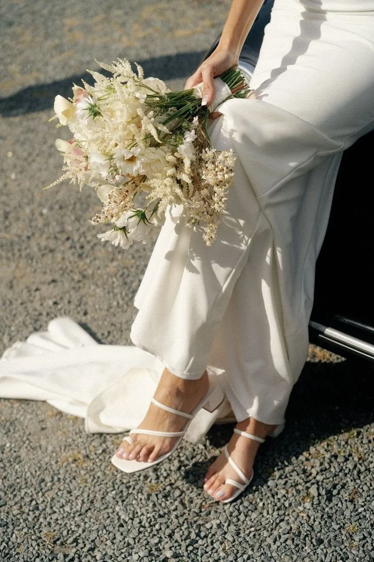 Close-up of a bride sitting on a black car, holding a bouquet of white and pastel flowers, wearing white wedding dress and high-heeled sandals, with a gravel ground.