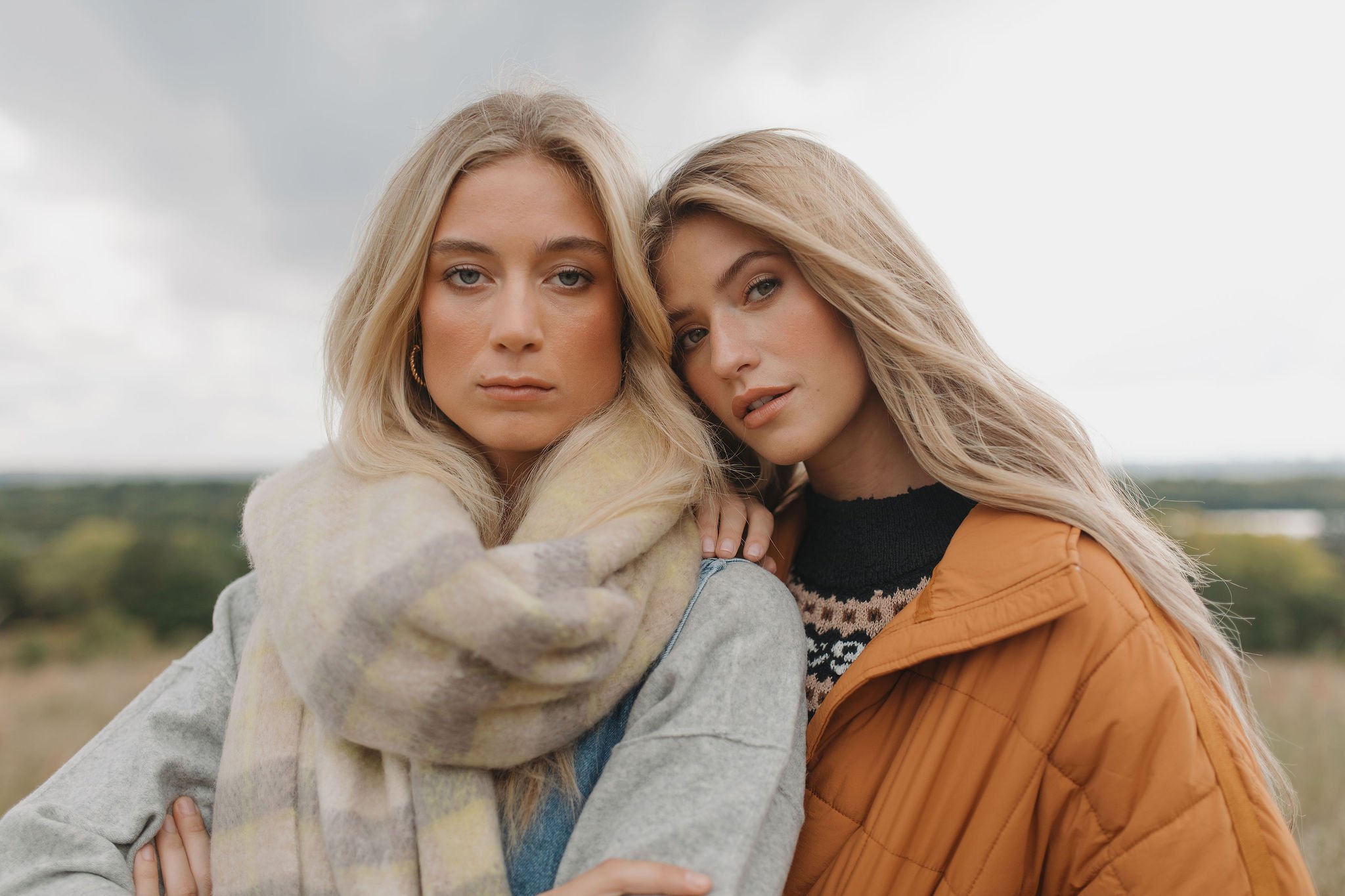 Two blonde women with fair skin posing outdoors against a cloudy sky, with one woman wrapping her arms around the other's shoulders.