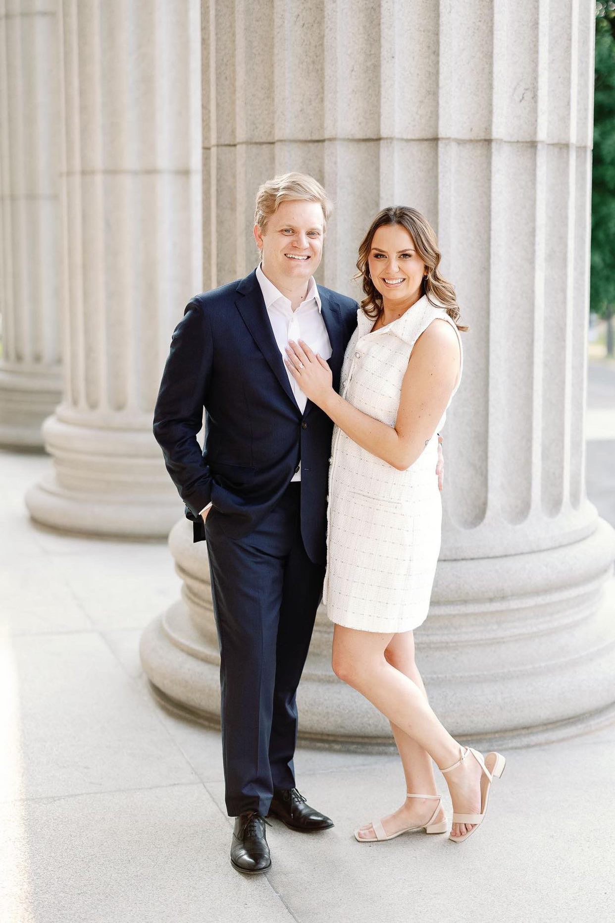 A smiling couple stands close together in front of large stone columns, with the woman resting her hand on the man's chest.