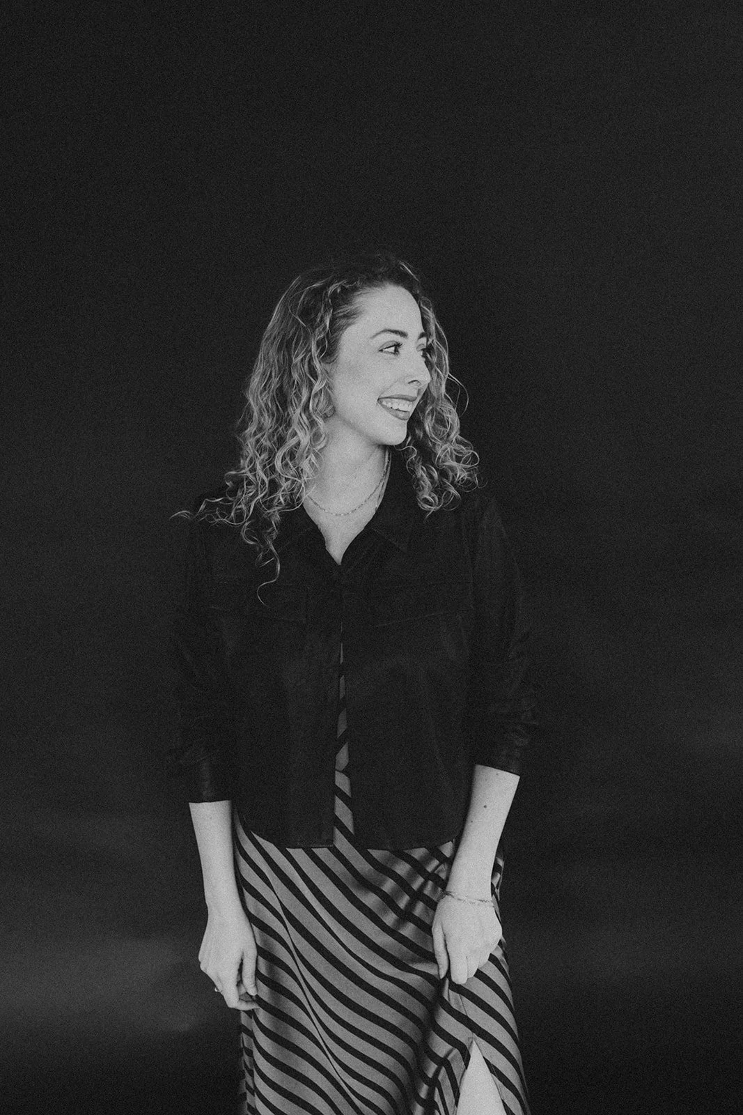 A woman with curly hair smiling and looking to her right, wearing a dark jacket and a striped skirt, standing against a dark background.