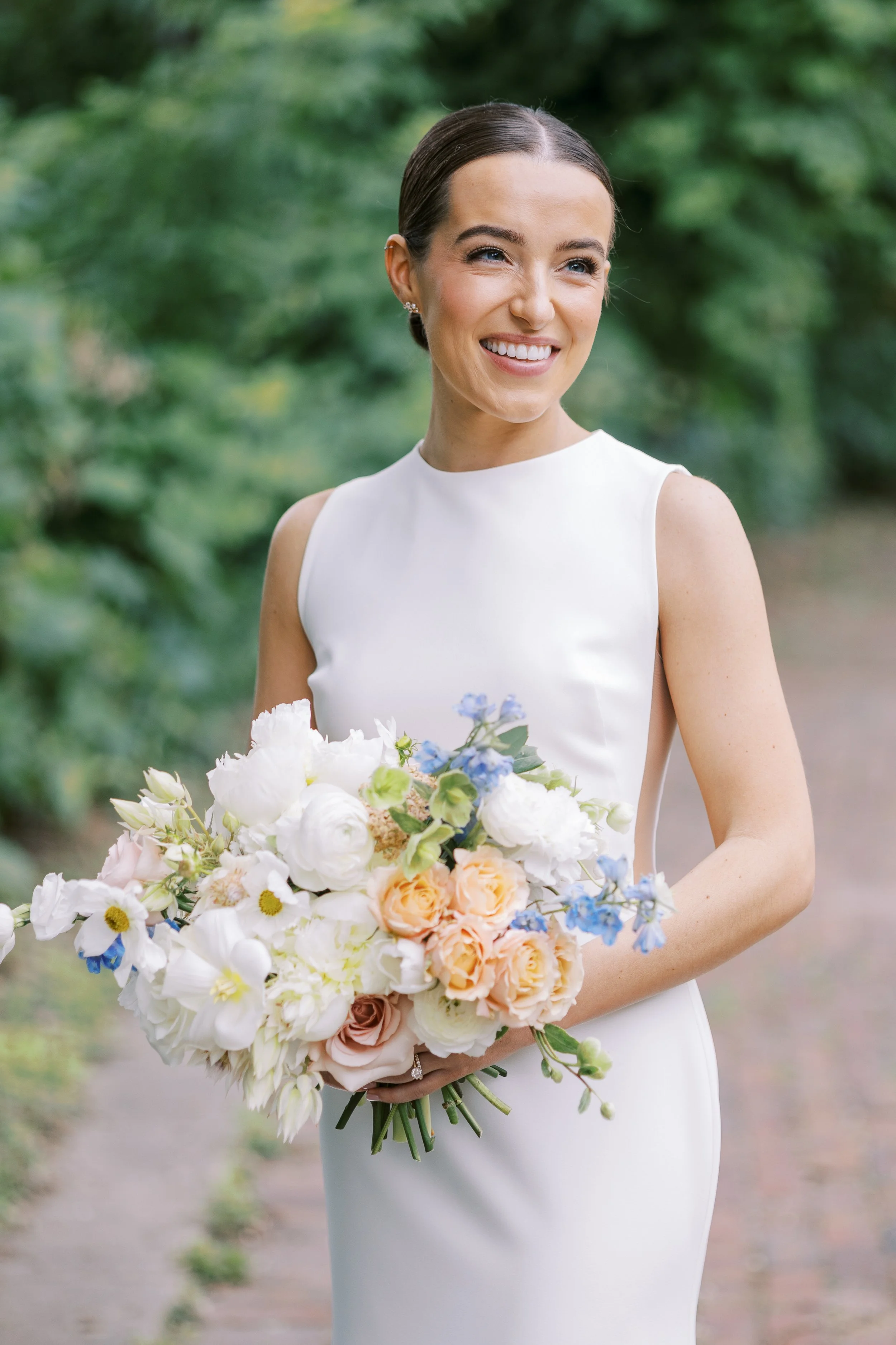 A smiling woman in a white dress holding a large bouquet of assorted flowers outdoors, with greenery in the background.