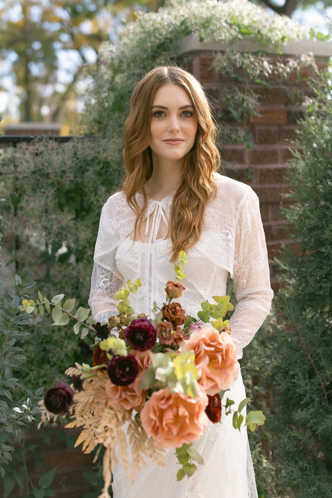 A young woman with wavy, light brown hair and blue eyes stands outdoors holding a bouquet of flowers. She is wearing a white lace dress with a tie at the neckline. The background includes greenery and a brick structure, with soft sunlight filtering through the trees.