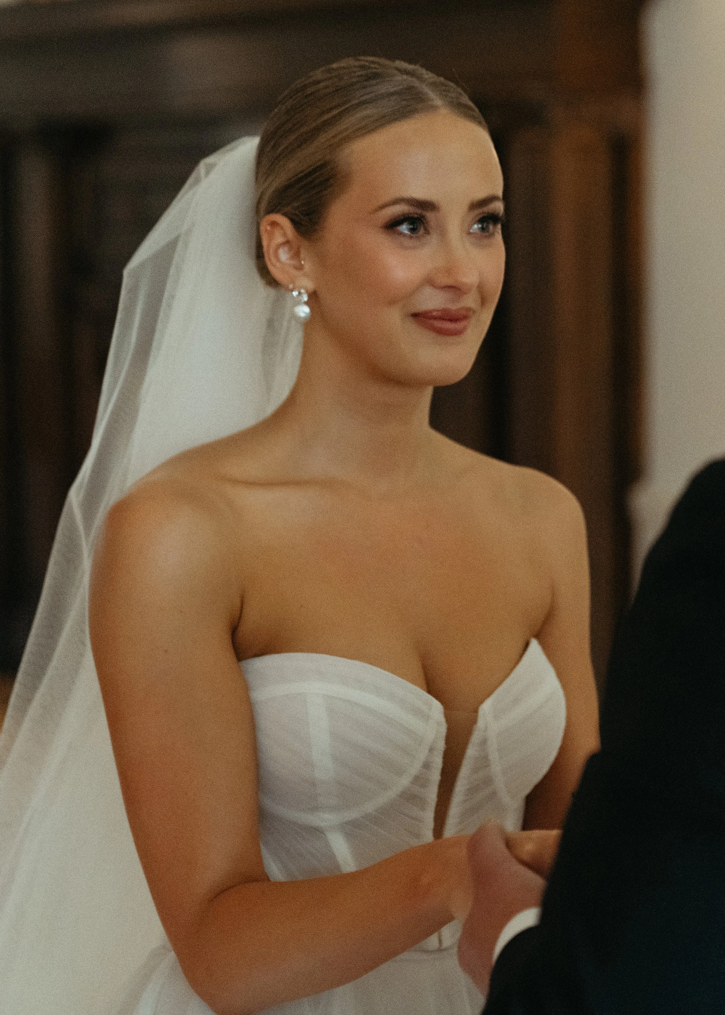 A bride with pixie-cut hair wearing pearl earrings and a strapless white wedding dress, smiling during a wedding ceremony.