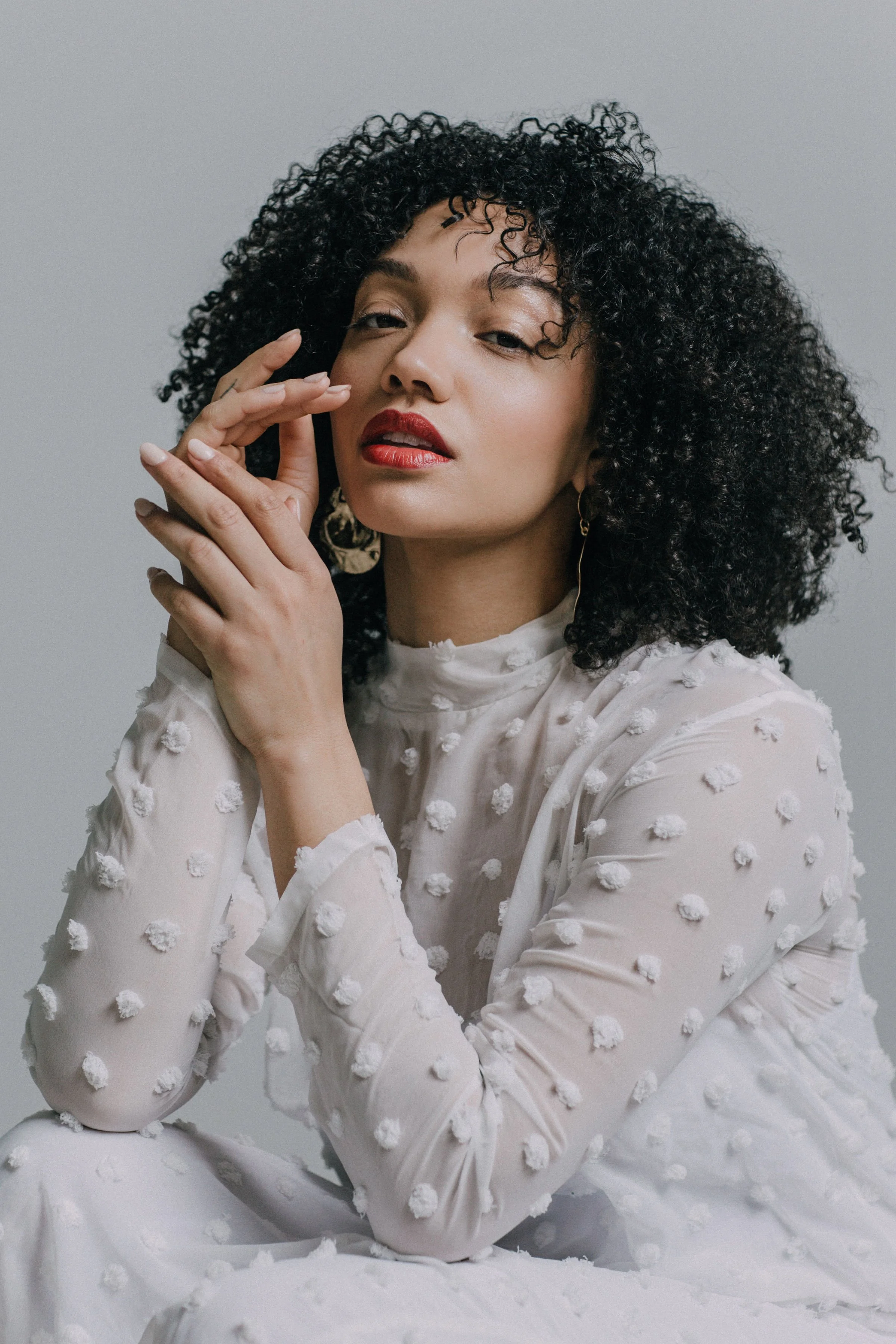 A woman with curly black hair and red lipstick wearing a white dress with textured polka dot pattern, posing with her hand near her face against a light gray background.