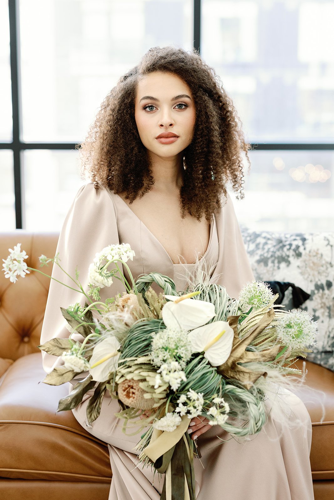A woman with curly hair holding a bouquet of white and green flowers, sitting on a tan sofa in front of large windows.