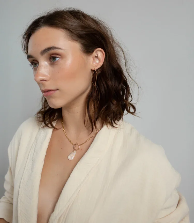 A woman with shoulder-length wavy brown hair, wearing a white top, gold geometric earrings, and a gold necklace with a large pearl pendant, standing against a plain light gray background.