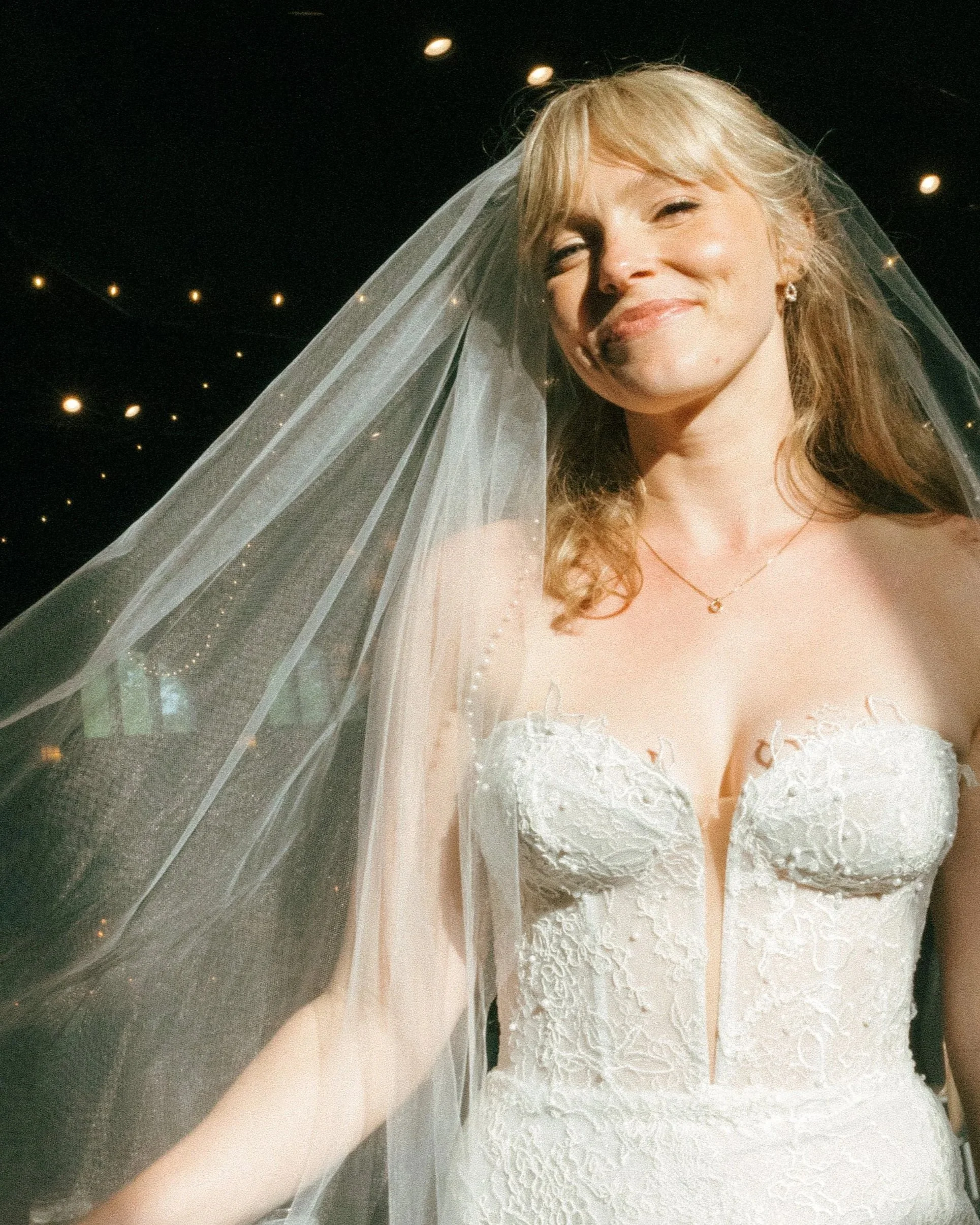 A smiling woman in a wedding dress with a veil, standing under a ceiling with small lights.