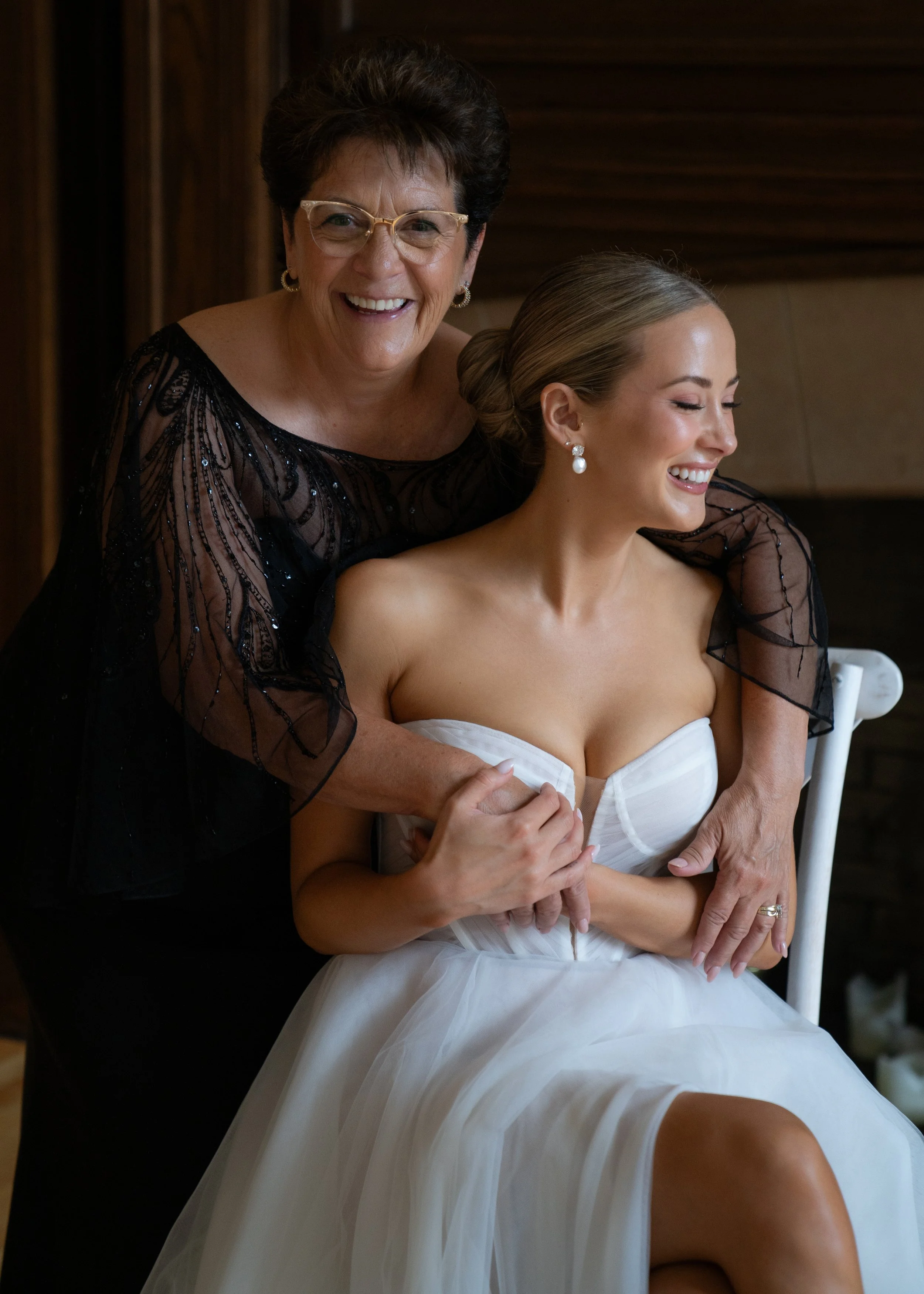 An older woman with short dark hair and glasses smiling, standing behind a younger woman in a strapless white wedding dress with her eyes closed, both wearing earrings, in a warmly lit indoor setting.