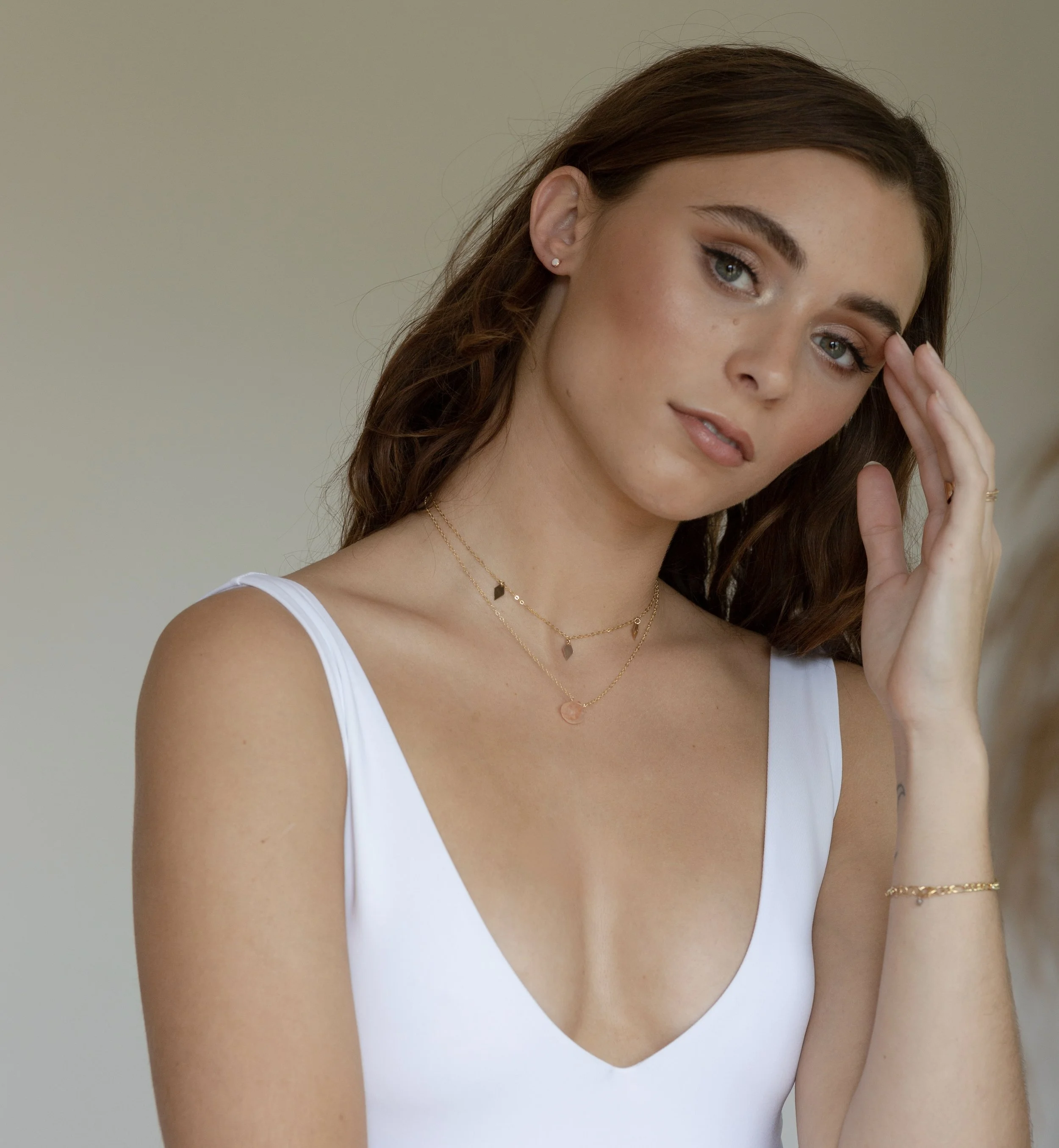 Close-up of a young woman with light skin and brown hair, wearing a white sleeveless top and gold jewelry, touching her temple with her right hand against a plain neutral background.