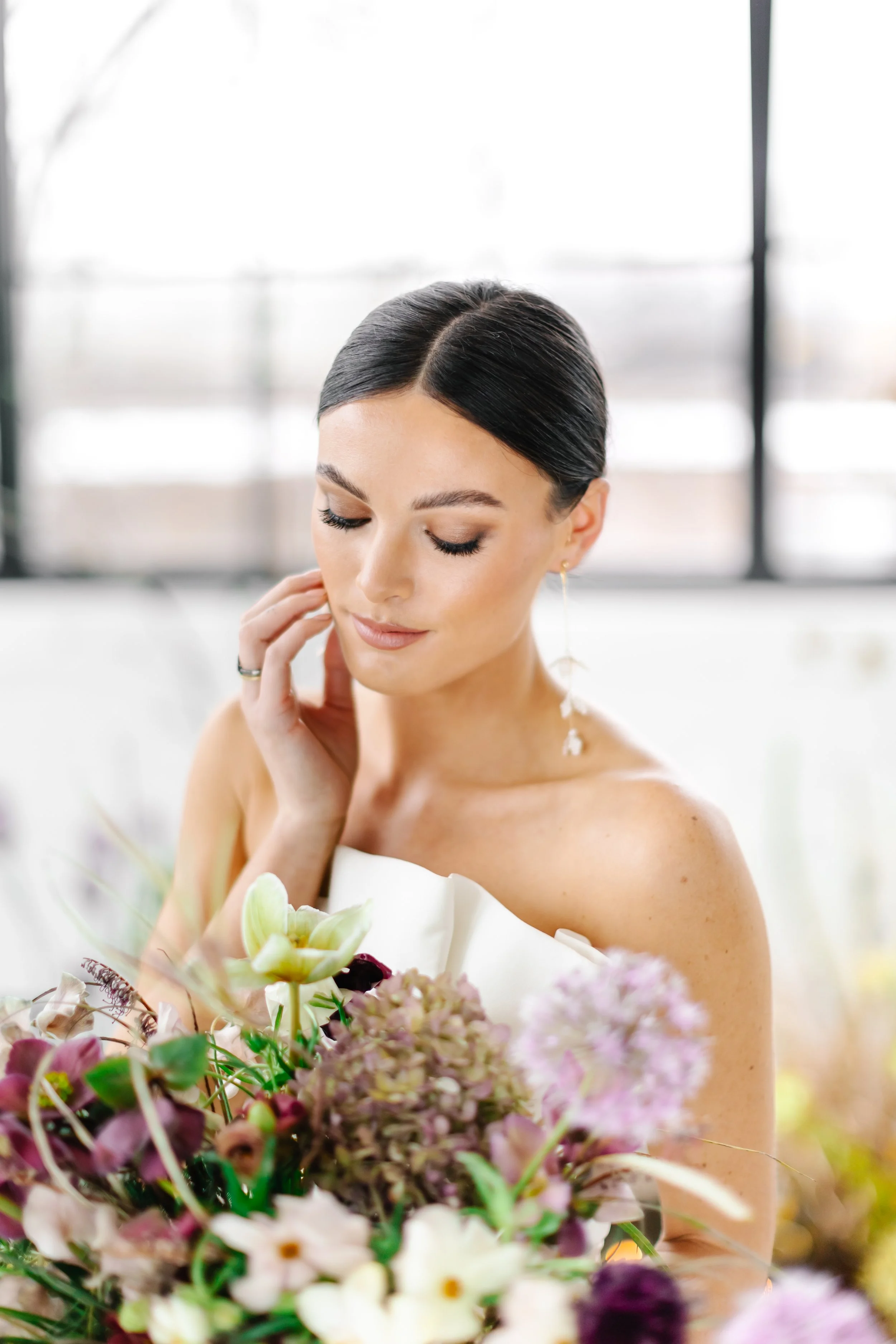 A woman with dark hair styled in an elegant updo, wearing a white strapless dress and earrings, looks down with a gentle smile at a bouquet of flowers in front of her. Background is bright and slightly blurred.