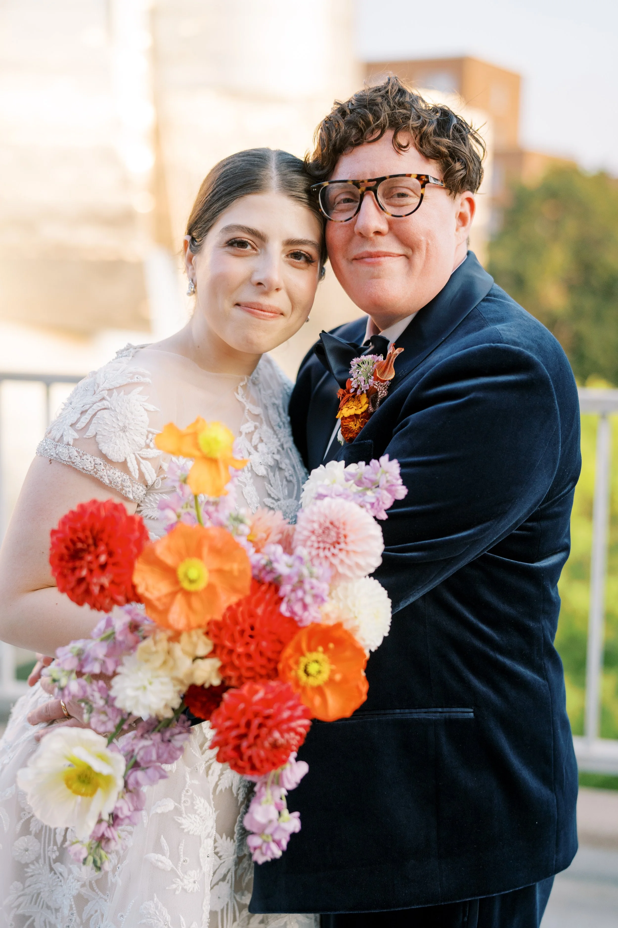 A bride and groom embracing, holding a colorful bouquet of flowers, outdoors on a sunny day.