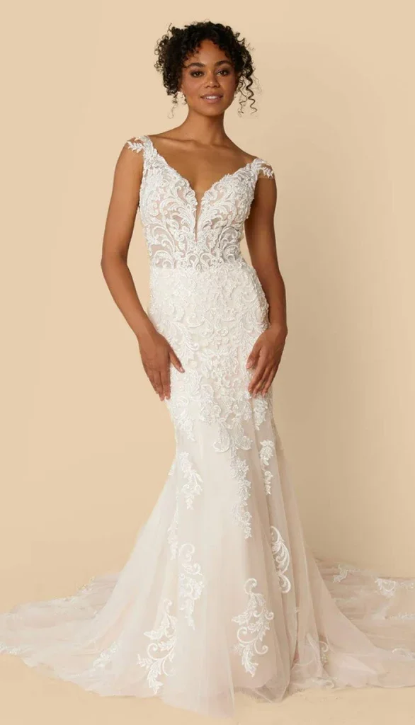 Woman in an elegant white lace wedding gown with a deep V neckline and off-the-shoulder sleeves, standing against a neutral background, with her hands gently resting at her sides.