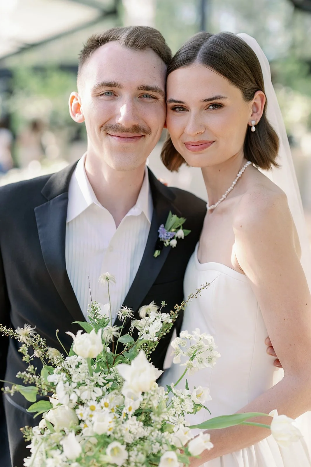A newlywed couple dressed in wedding attire, smiling and looking at the camera, outdoors with a background of trees and sunlight.