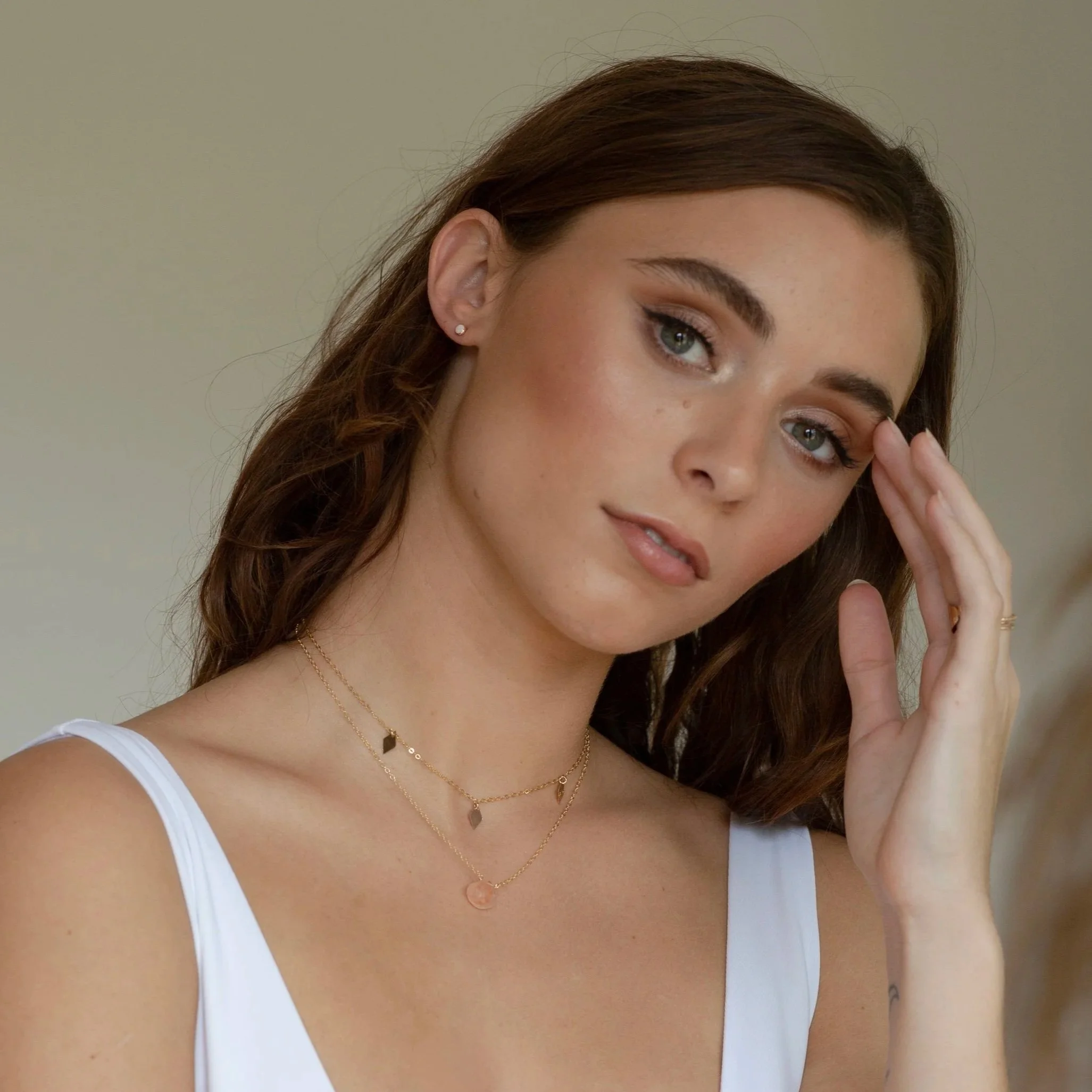 Close-up of a woman with reddish-brown hair, wearing a white top and layered gold necklaces, touching her face with a gentle expression.