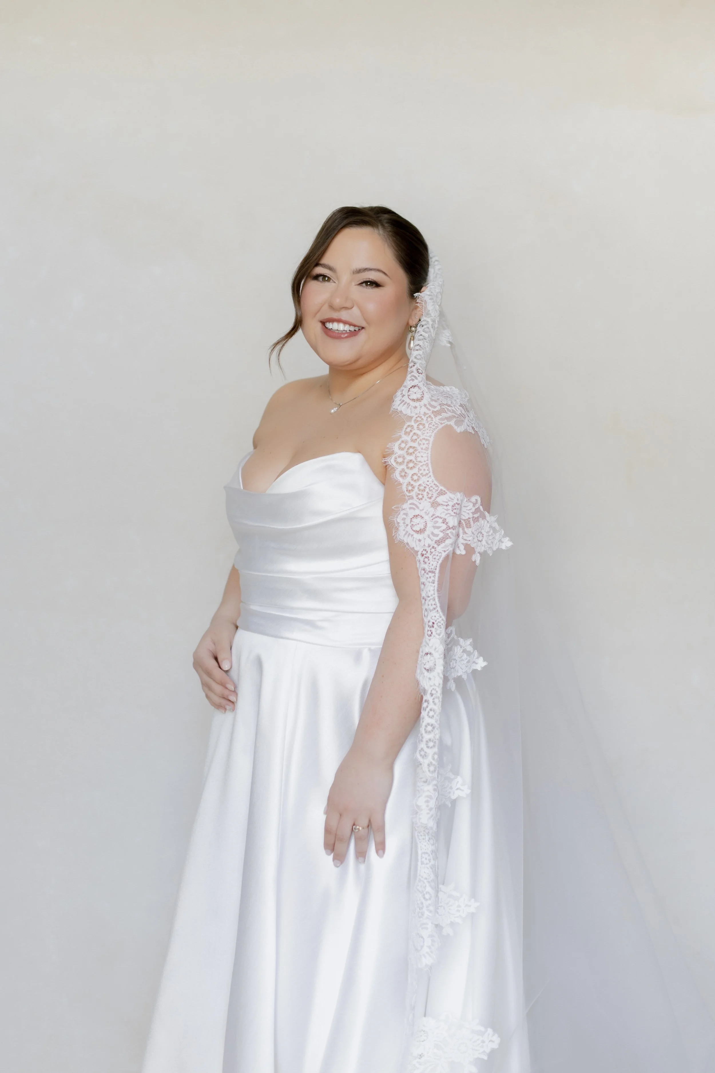 A woman in a white wedding dress with lace details on the shoulders and a lace veil smiling at the camera against a plain background.
