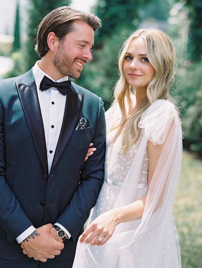 A smiling man and woman standing outdoors; the man wears a tuxedo and the woman wears a light-colored dress with sheer, puffed sleeves.