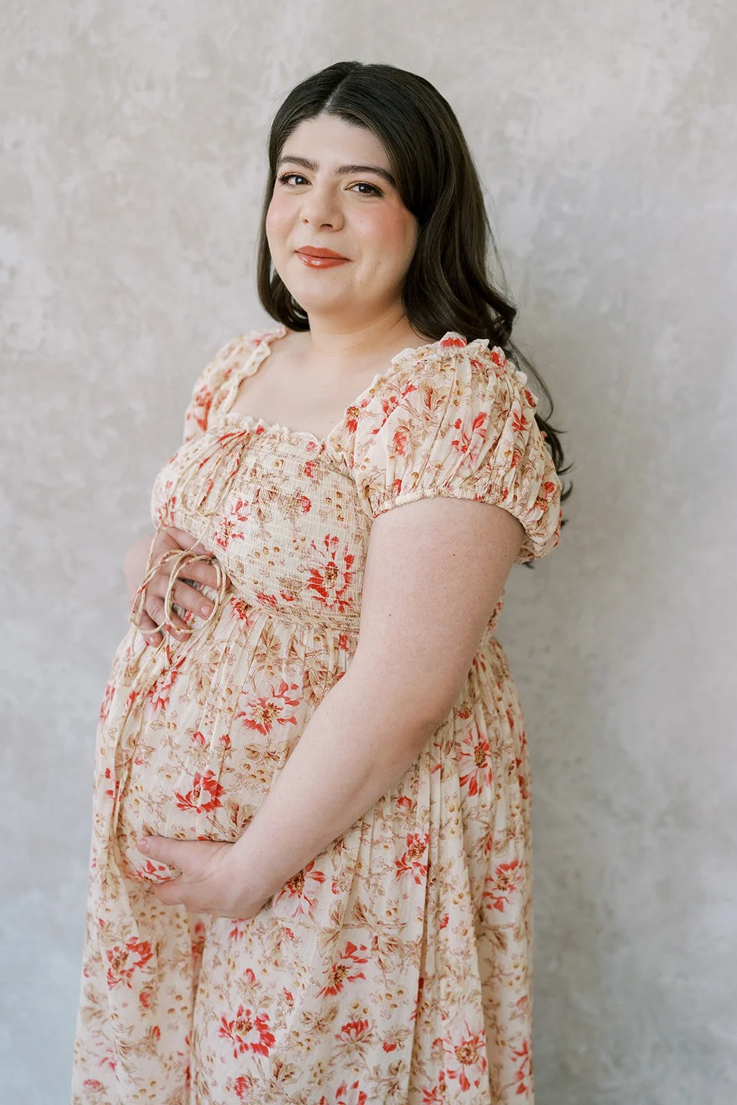 A pregnant woman with dark hair styled in soft waves, wearing a light floral dress with puffed sleeves, standing against a neutral textured background, gently holding her pregnant belly with one hand and smiling softly.