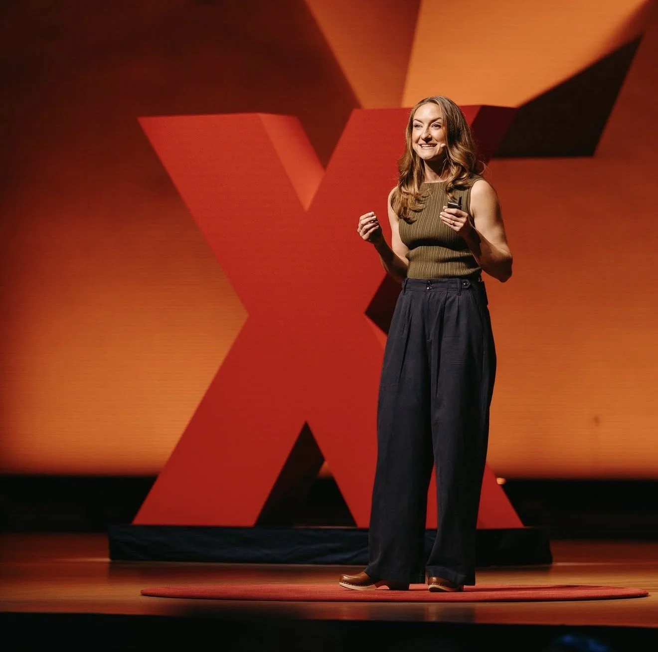 A woman giving a presentation on a stage with a large red 'X' in the background.