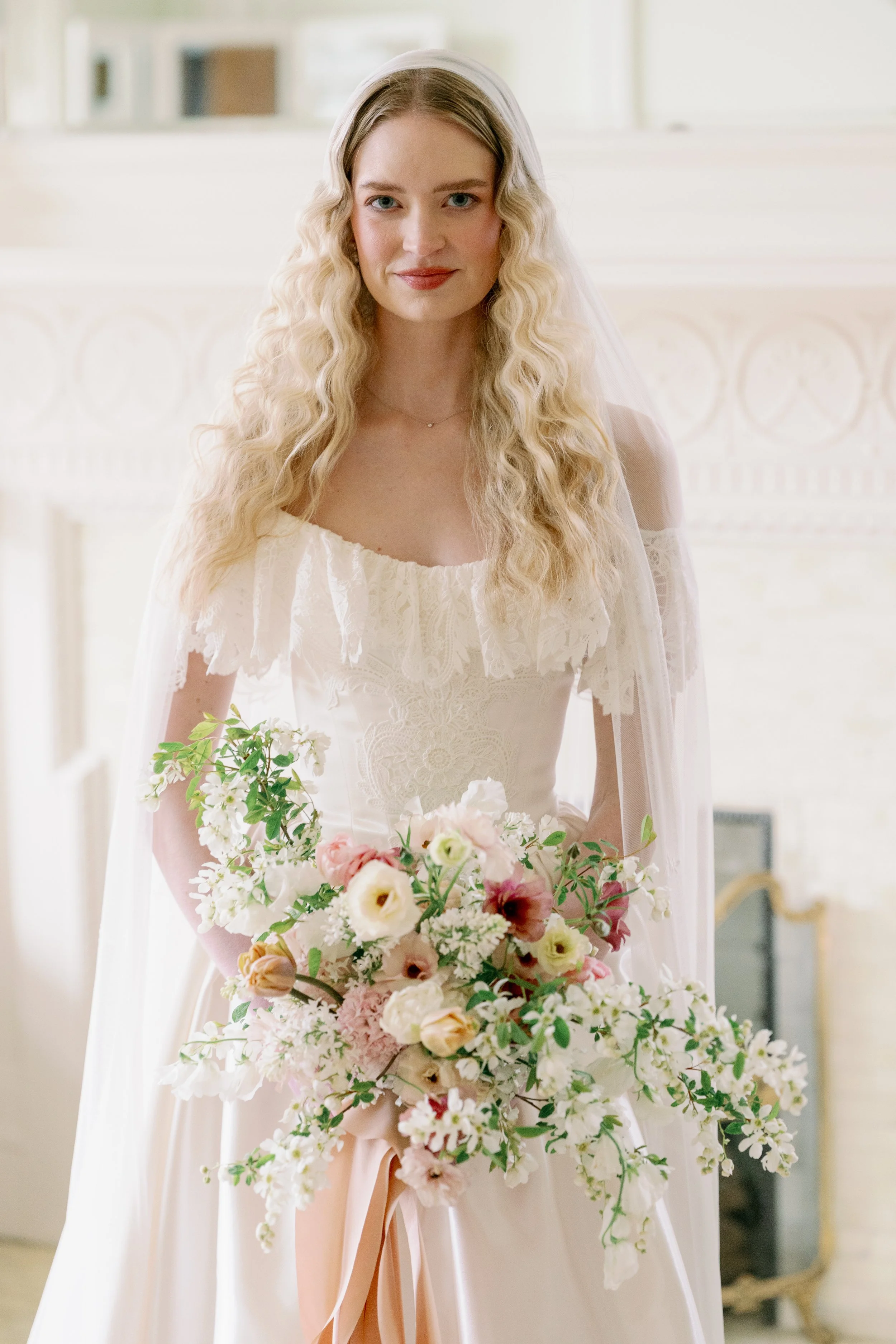 A woman with wavy blonde hair, wearing a white wedding dress and veil, holding a large bouquet of pink, white, and peach flowers.