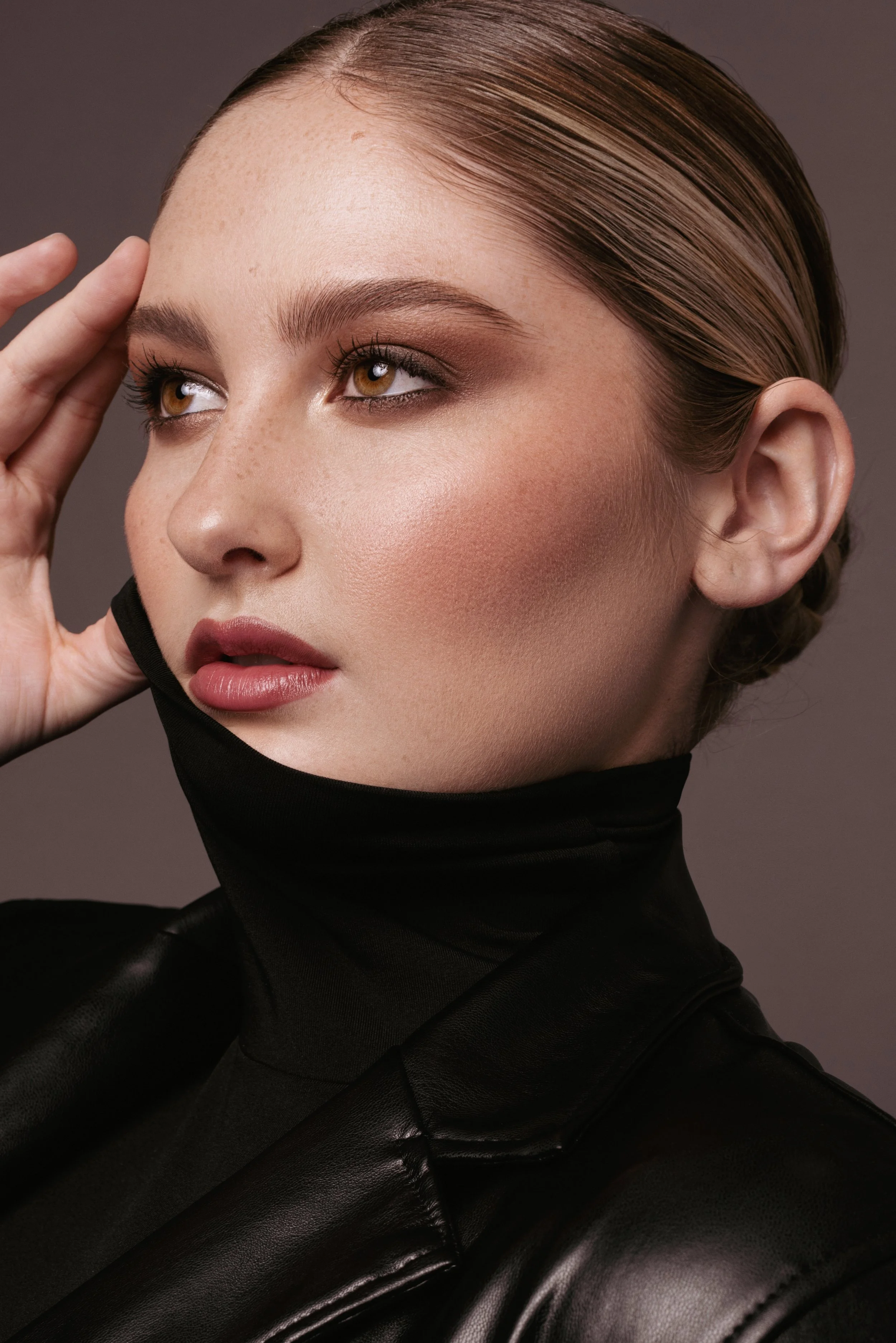 Close-up of a woman with brown hair, hazel eyes, and natural makeup, wearing a black turtleneck and touching her forehead with her hand.