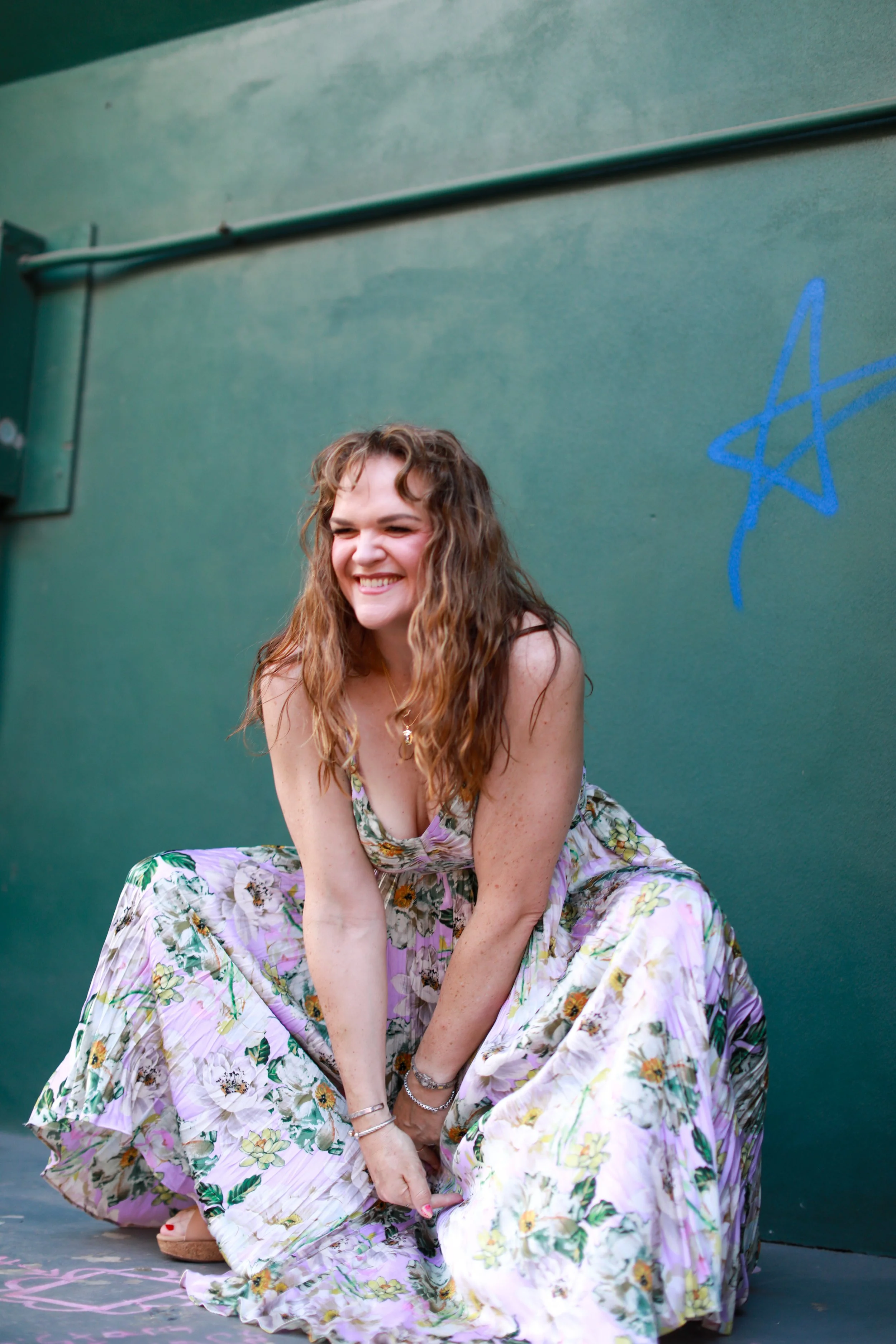A young woman with curly brown hair smiling, wearing a floral dress with pink, purple, yellow flowers, crouching against a green wall with blue graffiti, and chalk drawings on the ground.